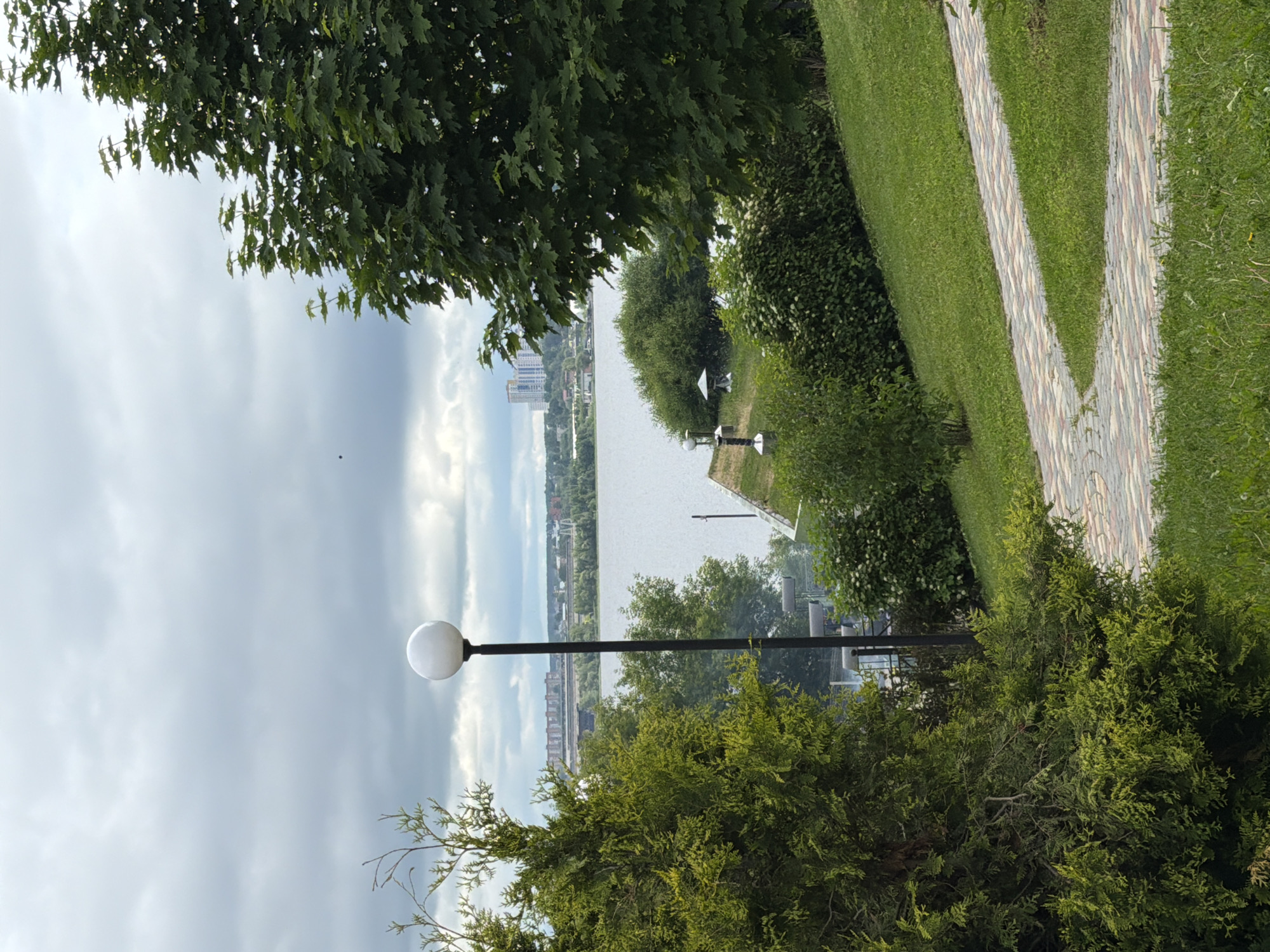 The image presents a daytime outdoor scene under an overcast sky, depicting a lush green park area overlooking a large body of water and a distant city.
In the immediate foreground, to the left, a dense, vibrant green coniferous bush or small tree (possibly a juniper or thuja) occupies a significant portion of the frame. To the right, a winding paved path, constructed from multi-colored interlocking bricks in shades of red, grey, and beige, traverses a grassy slope. The grass appears well-maintained.
Centrally located in the midground, a tall, slender black lamppost with a spherical white light fixture stands prominently. To its right, a large deciduous tree with numerous green leaves partially frames the scene from the upper right. Further into the midground, beyond the paved path and foreground vegetation, more green bushes and smaller trees line the edge of a wide, calm body of water, which reflects the grey sky. Along the near bank of the water, several smaller, darker ornamental lampposts or decorative bollards are visible, alongside a small, light-colored structure, possibly a gazebo or pavilion.
In the background, across the expanse of water, a cityscape is visible. It includes a cluster of modern, high-rise residential buildings and other urban structures. A long, linear structure, resembling a bridge or dam, spans the water horizontally in the middle distance, connecting to the city on the far shore. The sky above is entirely covered with light to medium grey clouds, indicating an overcast day. A tiny dark speck, potentially an insect or bird, is visible in the upper left portion of the sky.
This scene is identified as a view from the Izhevsk Embankment (Набережная Ижевского пруда) in the city of Izhevsk, Russia, looking across the Izhevsk Pond towards the urban developments on the opposite bank, likely encompassing parts of the city and the Izhevsk Dam.