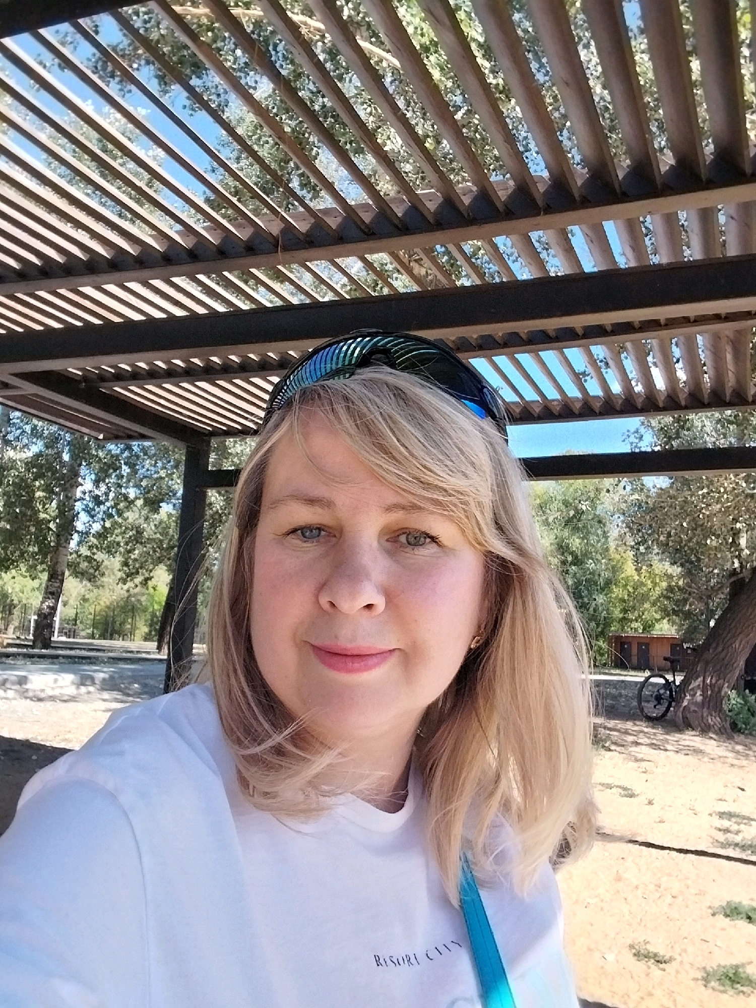 A woman with blonde hair and blue eyes smiles as she takes a selfie under a wooden pergola. She is wearing a white t-shirt and sunglasses on her head. In the background, trees and a small building are visible. The image appears to have been taken outdoors in a park or recreational area.