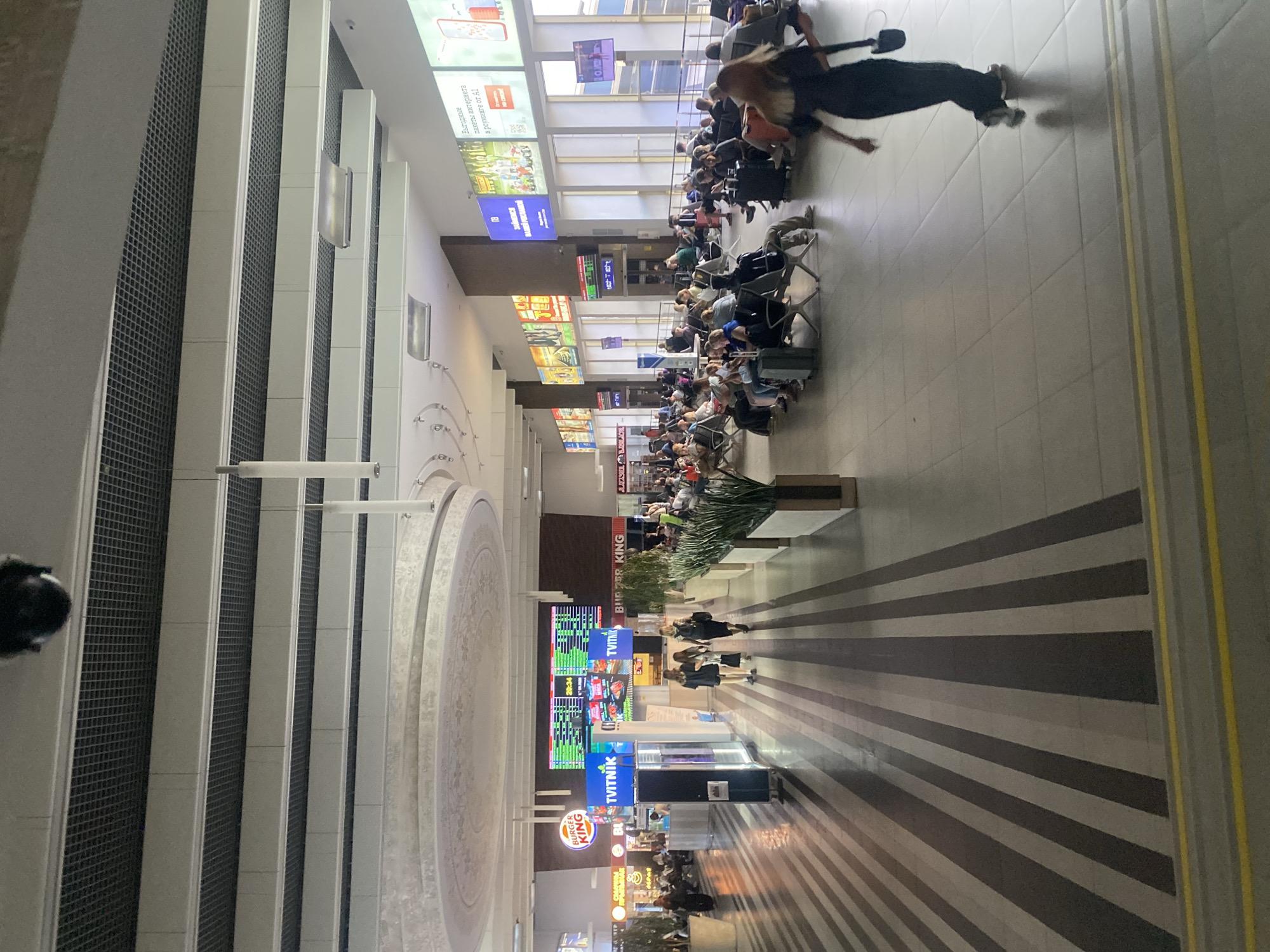 This is a photograph taken inside a modern, spacious building, likely a transportation hub such as an airport or train station, located in Minsk, Belarus. The architecture features a high ceiling with suspended lighting fixtures and a grid-like ceiling panel system. A black security camera is visible in the upper left corner.
The floor is covered in large square tiles and features a distinctive pattern of alternating dark gray and lighter gray stripes. The walls are adorned with large digital displays and advertisements. One prominent display shows a timetable with green and red entries, likely indicating departure or arrival information. Other advertisements feature vibrant images of food and products.
In the foreground, a woman in a dark dress and white sneakers walks away from the camera. Several people are seated on benches in the middle ground, waiting, many with luggage at their feet. These individuals appear to be of various ages and backgrounds. In the left mid-ground, there are retail or food establishments, including a Burger King. Green potted plants are strategically placed throughout the area, adding a touch of nature to the interior. The overall atmosphere suggests a busy but orderly public space.