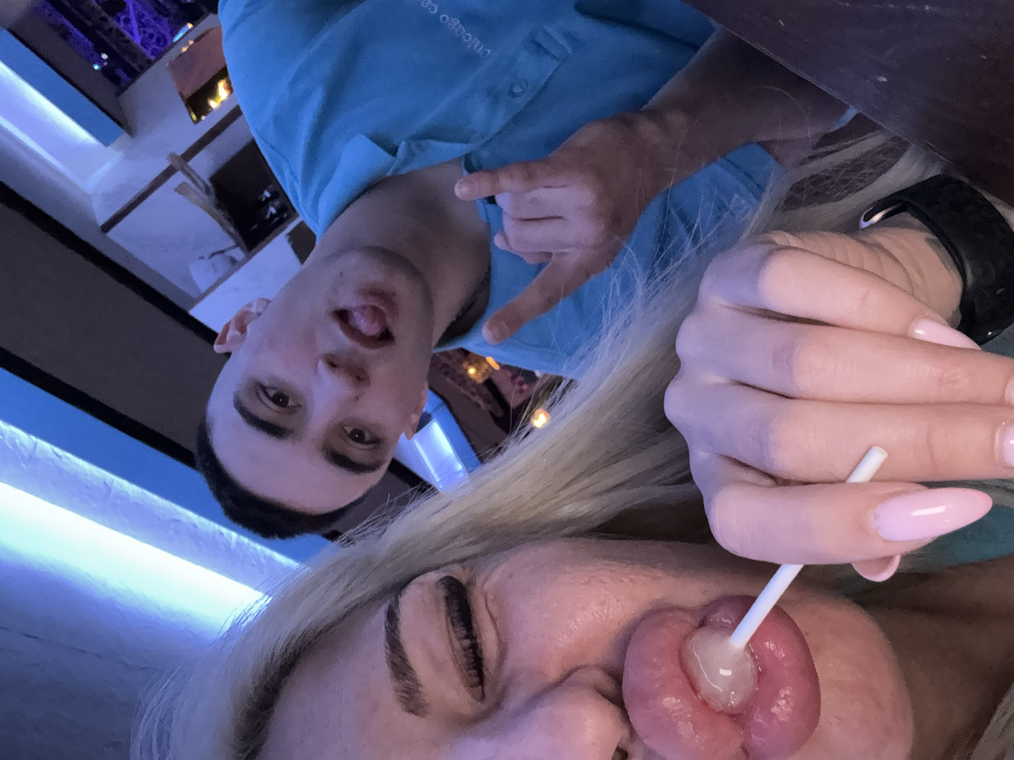 A selfie-style image, rotated counterclockwise, depicts a man and a woman in an indoor setting, potentially a restaurant.
The woman is in the foreground, her face partially visible. Her eyes are closed and she has long blonde hair. She is holding a lollipop, with the candy part touching her tongue. Her hand is visible, featuring manicured pink nails. A black smartwatch is on her wrist.
The man is positioned behind and to the left of the woman. He is making a "hang loose" hand gesture. He has short dark hair. He is wearing a light blue collared shirt with text that includes "chicago".
The environment includes blue lighting. A shelf with various items is visible in the background.
It's not possible to determine whether the location is in Plovdiv, Bulgaria from the image alone.