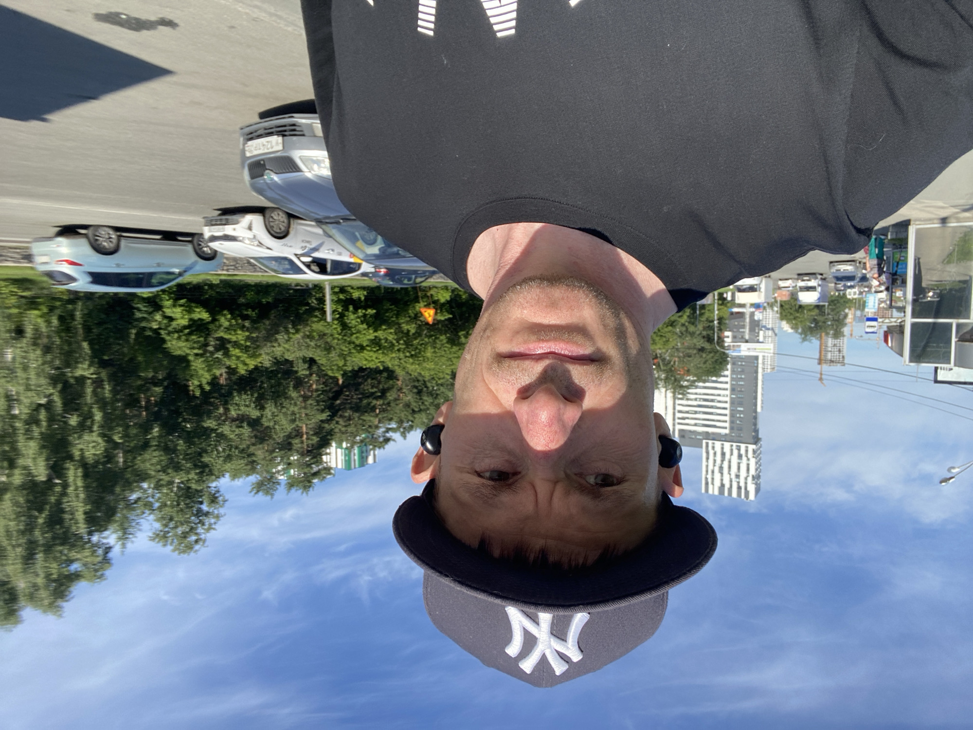 The image shows a man wearing a black t-shirt and a New York Yankees baseball cap, with black wireless earbuds in both ears. He is taking a selfie outdoors, with a city street and buildings in the background. The street has several cars, including white taxis and a light blue SUV. Trees and a partly cloudy blue sky are also visible. The overall impression is that of a casual, sunny day in an urban environment.
Based on visual cues, the location is likely Yekaterinburg, Russia. The architecture of the buildings, the style of the license plates on the cars (e.g., the visible plate on the grey car reads "H 124 TP 96"), and the presence of taxis with the "Yandex.Taxi" logo are consistent with this location. The building with the distinctive vertical white and grey pattern in the background is identifiable as the "Skyline" residential complex, a prominent landmark in Yekaterinburg.