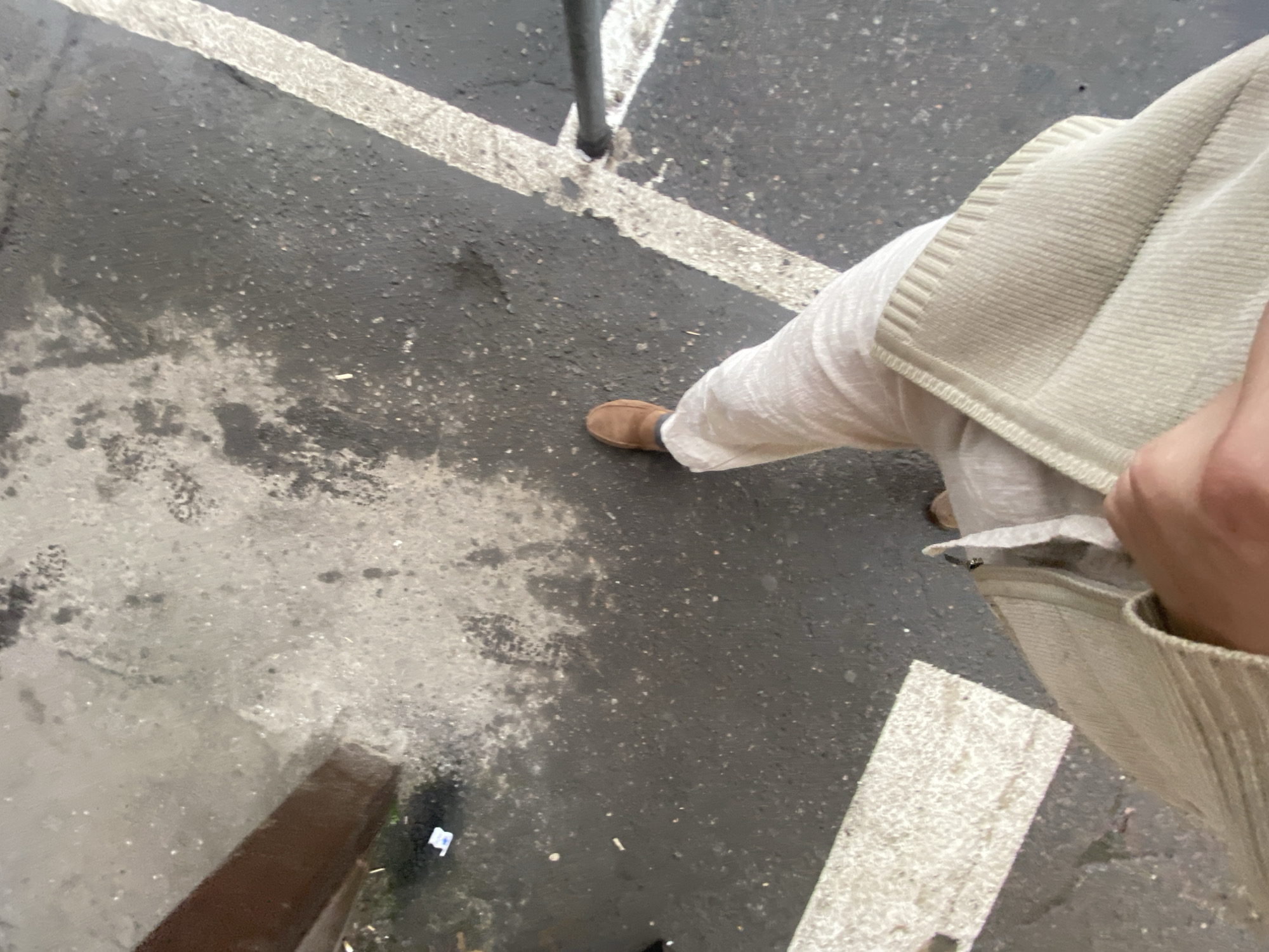 The image shows a ground-level view of a person's legs and feet, standing on a wet, asphalt surface marked with white lines. The person is wearing light-colored trousers and brown loafers. In the foreground, a hand is visible, holding onto a light-colored jacket. To the left of the frame, a dark brown, horizontal beam is visible, and behind it, some greenery and a small piece of white paper. The environment appears to be an outdoor urban setting. No specific details or landmarks are visible to indicate the location within Moscow, Russia.