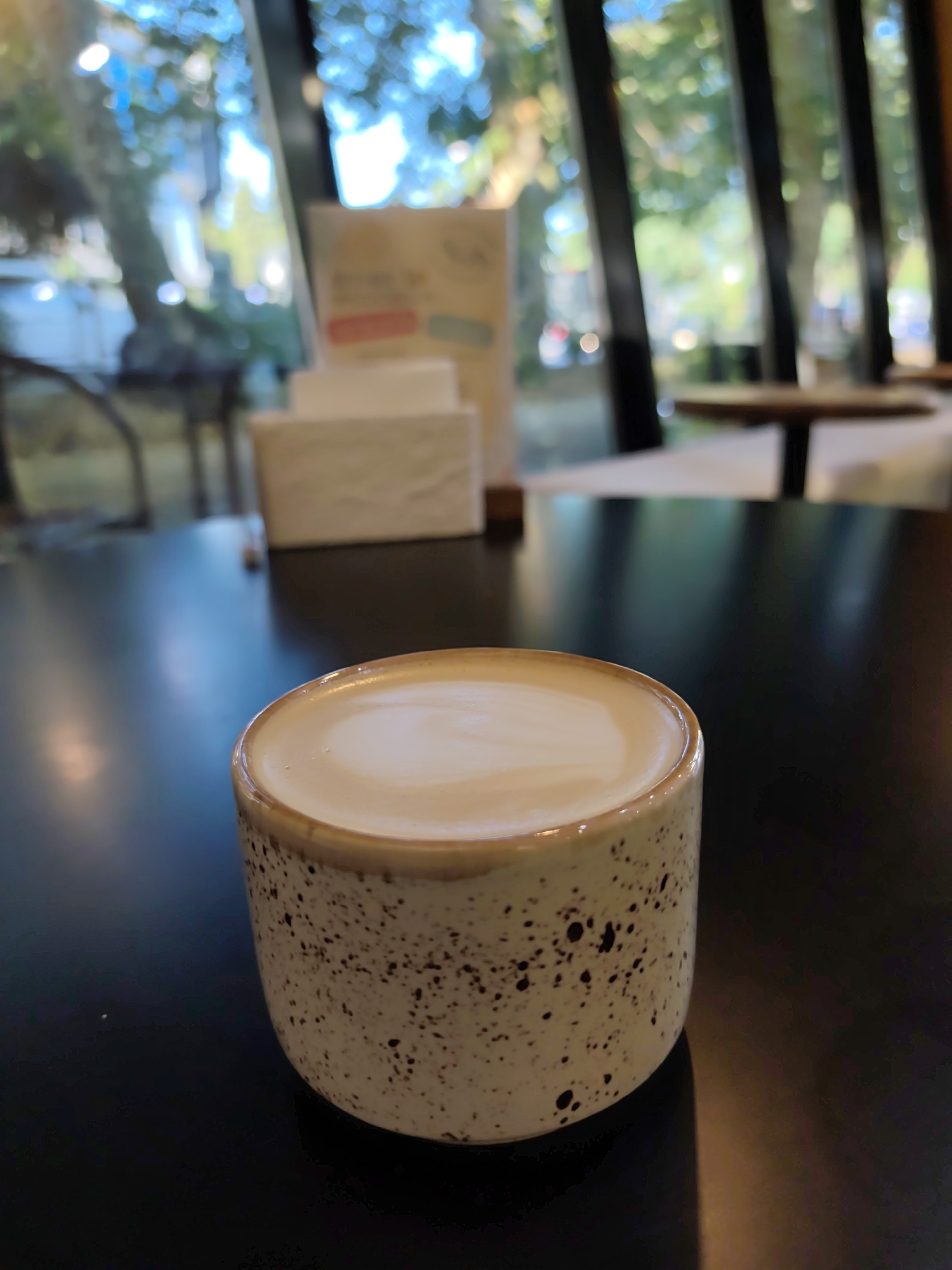 An overhead shot captures a close-up of a speckled ceramic cup filled with latte art, placed on a dark, reflective table. The cup's brown rim complements the speckled pattern of dark spots on a light background. The background is a blur of greenery and lights, suggesting an outdoor or well-lit indoor setting with large windows. Out-of-focus tables and chairs are visible in the distance, hinting at a cafe or restaurant environment. The overall impression is one of quiet contemplation, possibly in a cafe in Sochi, Russia, where the natural light filtering through the windows creates a serene ambiance.