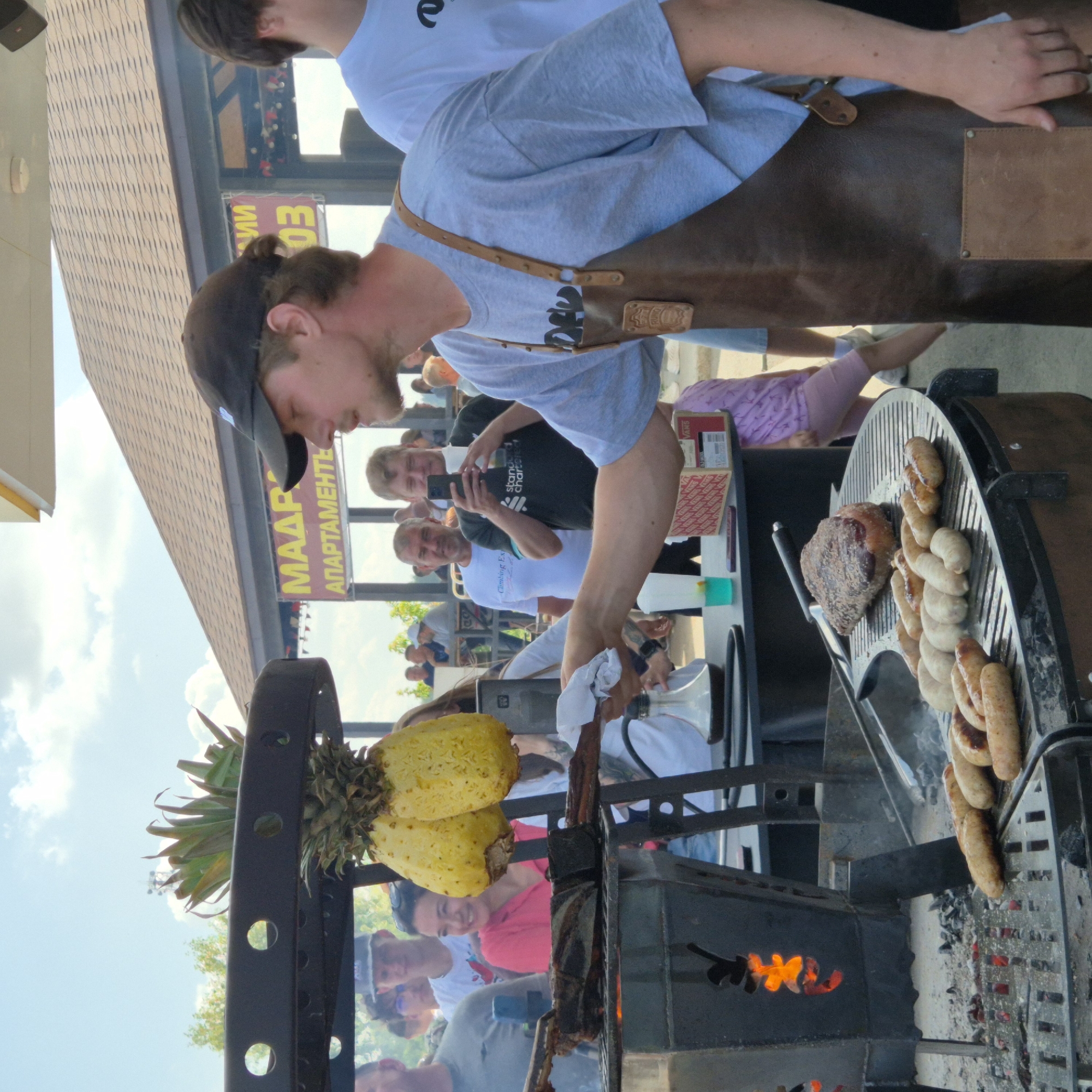 This image depicts an outdoor grilling event, likely in the city of Promyshlennaya, Russia. In the foreground, a man wearing a light blue t-shirt and a brown leather apron is standing beside a large grill. He appears to be actively involved in the cooking process, holding a pair of tongs. The grill itself is made of dark metal, with a raised grate holding several sausages and a large piece of meat. Flames are visible within the grill's firebox.
Hanging above the grill are two halves of a pineapple. A group of people are gathered in the background, observing the grilling. One woman in a pink shirt is smiling and looking towards the grill. Others are standing, talking, and some are holding phones. A banner with Cyrillic text and the number "03" is visible in the background, suggesting a business or event advertisement. The environment appears to be a public outdoor space with a clear sky and some trees visible. The overall scene conveys a festive and casual atmosphere centered around food preparation.