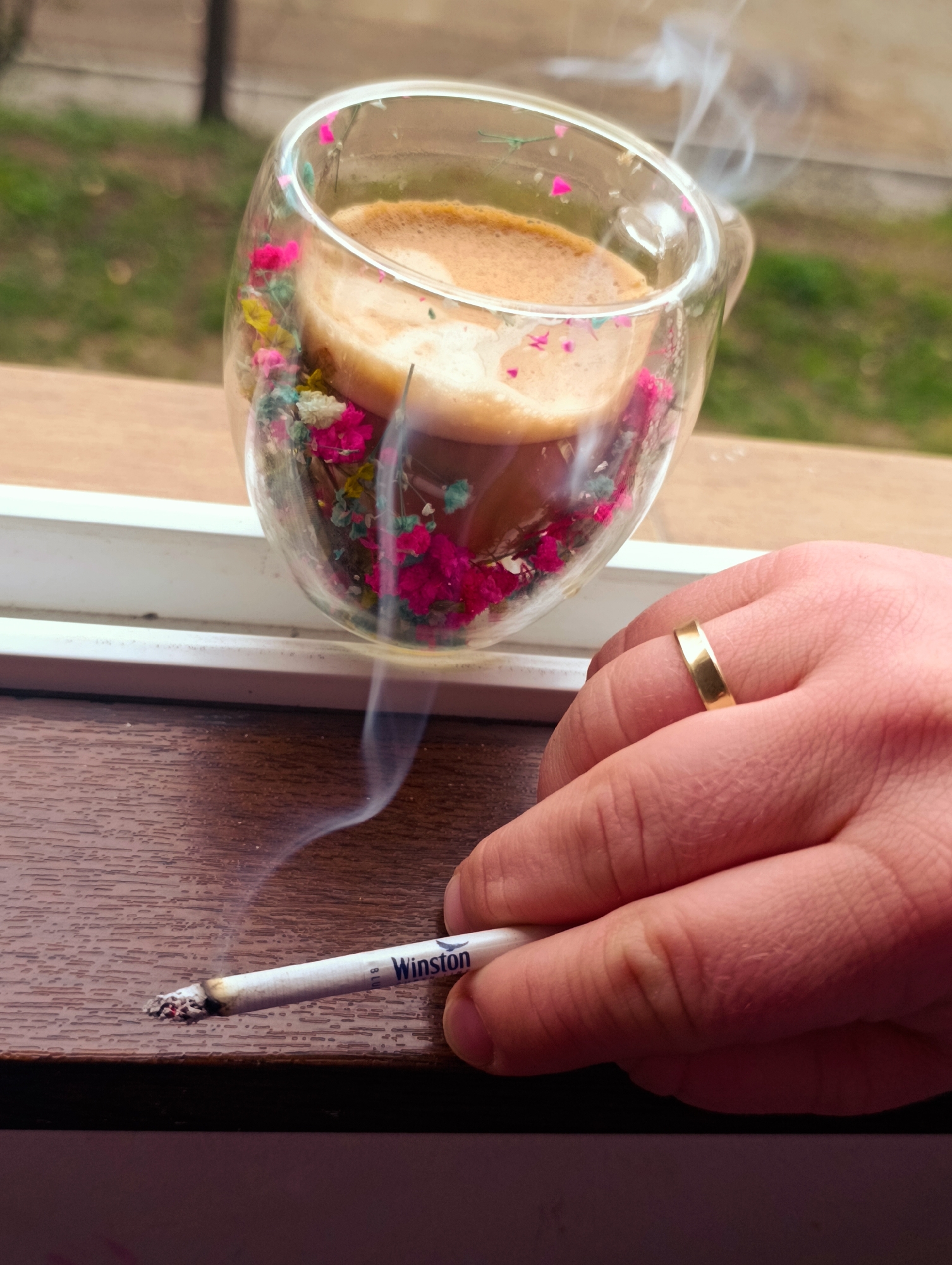 A photograph taken indoors in an unknown location, possibly Kardzhali, Bulgaria, shows a lit Winston cigarette on a brown surface next to a hand wearing a gold wedding band. A double-walled glass mug filled with coffee and decorated with small, dried flowers sits on a white ledge above the cigarette. Smoke rises from both the cigarette and the mug. A blurry view of greenery is visible through the window behind the mug.