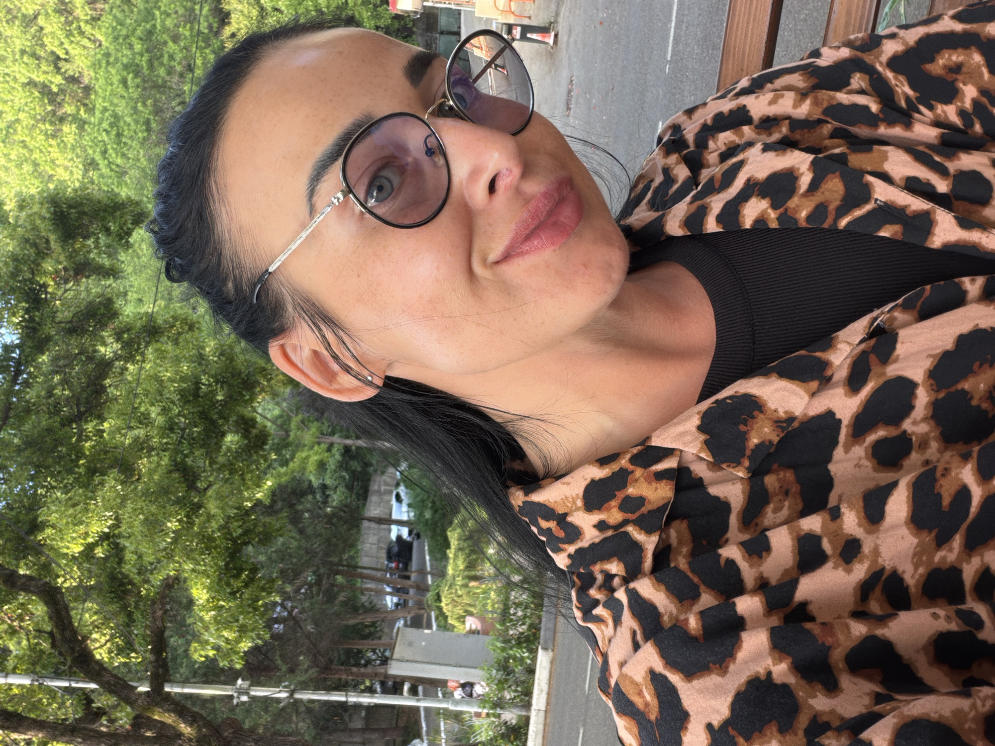 This is a close-up, portrait-style, outdoor photograph of a woman wearing round, wire-rimmed glasses. She has dark hair pulled back and is wearing a black top beneath a leopard-print shirt. Her head is tilted and she is looking towards the camera with a slight smile. The background is out of focus, but appears to be a green, leafy area, possibly a park or garden, with some trees and sunlight filtering through. There are some hints of a paved area and structures in the distance, suggesting an urban or semi-urban setting. The lighting suggests it is daytime, likely sunny given the brightness and shadows. The overall mood is casual and relaxed. There is no visible text in the image. The location context is Sochi, Russia.