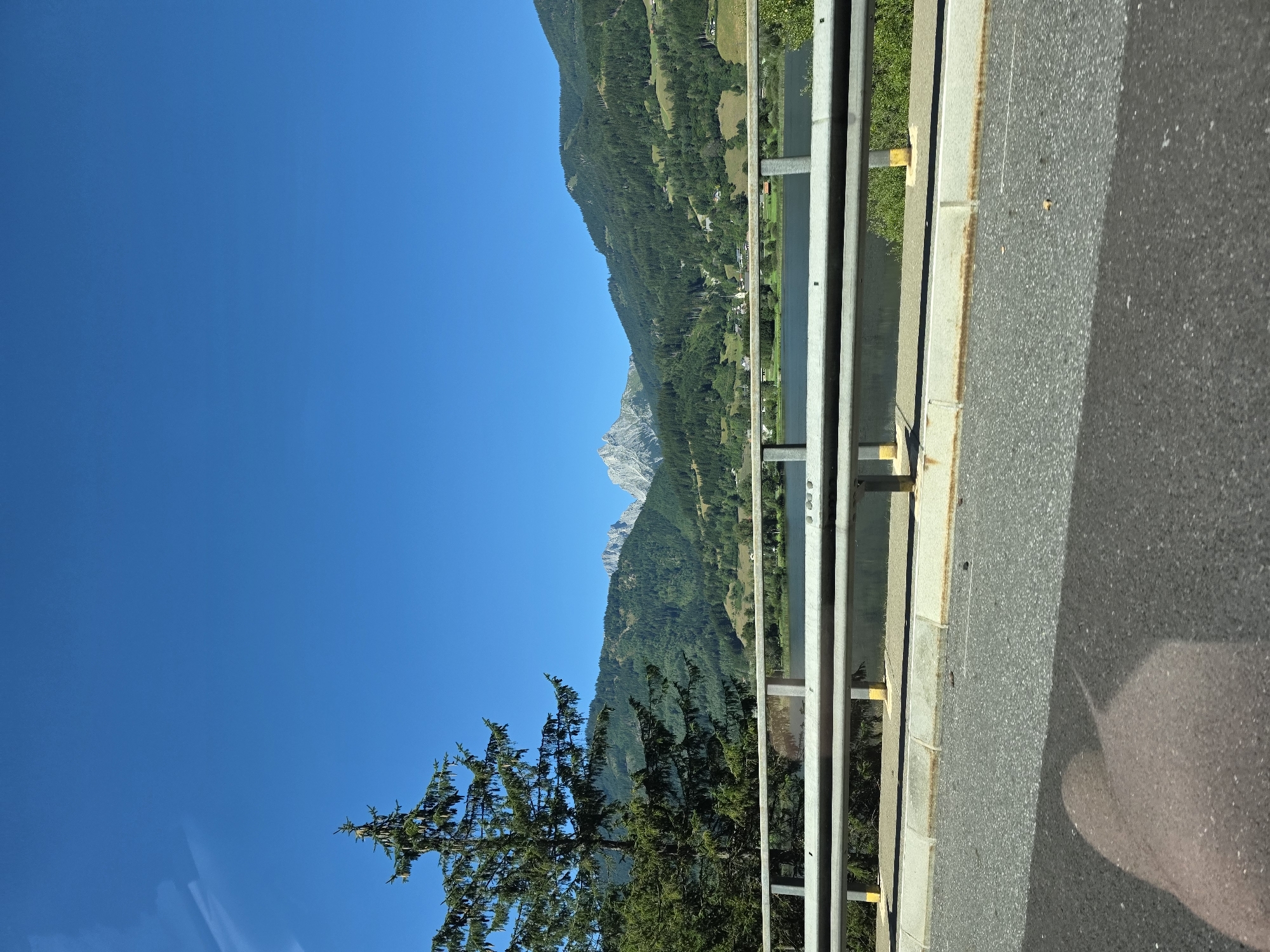 The image depicts a scenic view of a mountainous landscape with a clear blue sky. In the foreground, a metal guardrail runs horizontally across the frame, separating a paved road from a body of water. A large evergreen tree is visible on the left side of the image, with its branches extending upwards. Beyond the guardrail and the water, rolling hills covered in dense green forests lead up to jagged, rocky mountain peaks in the distance. A small village or settlement with scattered buildings can be observed nestled among the trees on the hillsides. The overall impression is of a tranquil, natural environment. Based on the provided context, this scene is located in Au bei Gaishorn am See, Austria.