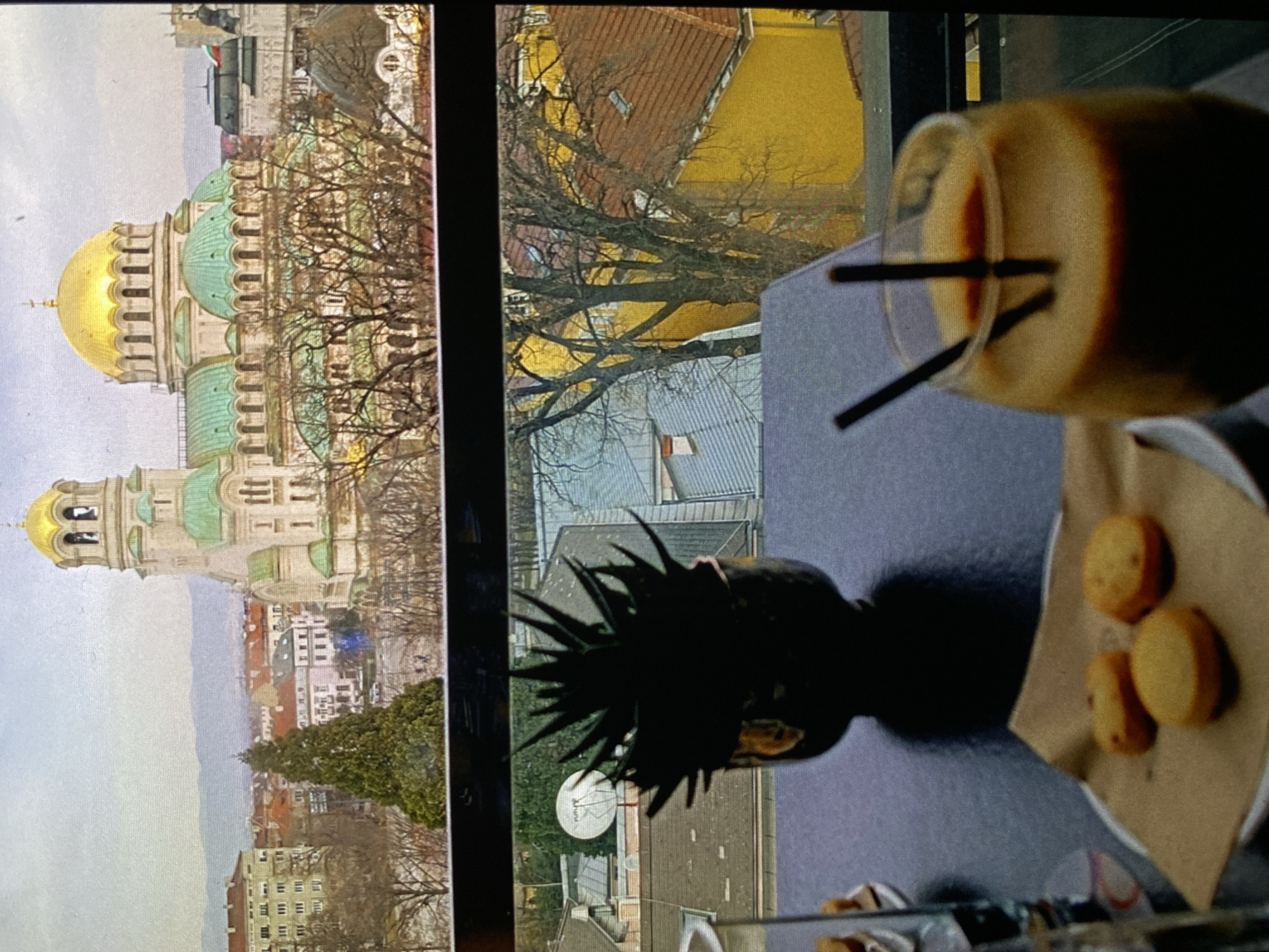 The image displays a scene viewed from indoors, looking out through a window onto a cityscape. On the right, occupying the foreground, a dark table holds several items. A clear glass contains a light brown beverage with two dark straws. Next to it, a small, dark, spiky decorative plant sits in a pot. Further right, a white plate holds three round, golden-brown snacks, possibly cookies or small cakes, resting on a piece of brown paper.
Through the window, on the left side of the frame, the prominent structure is the Alexander Nevsky Cathedral, a distinctive landmark in Sofia, Bulgaria. Its grand architecture is characterized by a large golden central dome and multiple smaller green-domed towers. Bare trees, suggesting late autumn or winter, partially obscure parts of the cathedral and the surrounding urban environment. The cityscape extends into the distance, featuring buildings with diverse roof colors, including red, brown, and yellow, and a satellite dish visible on one of the nearer rooftops. The sky above is overcast and grey, indicating a cloudy daytime.
The scene suggests a relaxed moment, enjoying refreshments indoors while observing the city view. No discernible text is visible. While the provided location context is Bankya, Bulgaria, the main landmark depicted is the Alexander Nevsky Cathedral located in Sofia.
