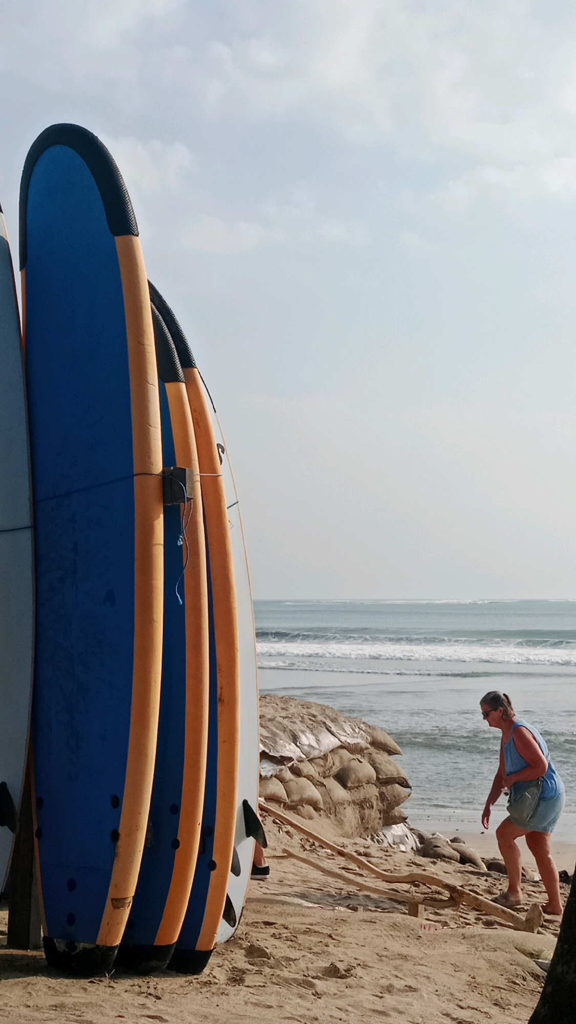An outdoor, vertical, medium shot captures a beach scene in Kuta, Indonesia. In the foreground, several surfboards are propped upright, with the leftmost one being a prominent blue and orange design. In the middle ground, a woman, wearing a blue denim dress and sunglasses, walks along the sandy shore. Behind her, sandbags are piled as a barrier, and the ocean with gentle waves stretches to the horizon under a pale sky. The lighting suggests it is daytime, likely mid-morning or late afternoon, casting soft shadows.