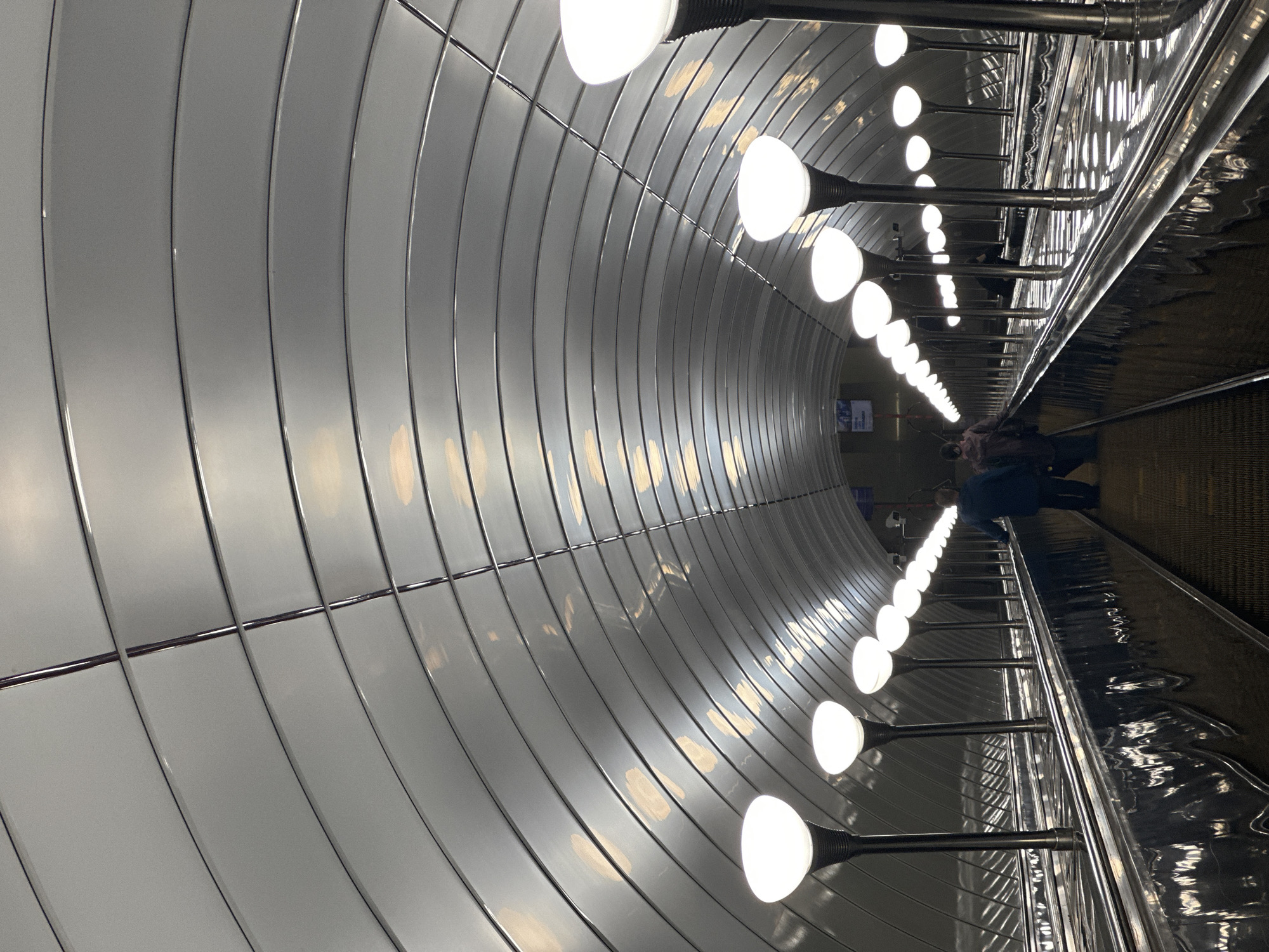 This image depicts an escalator in the Saint Petersburg Metro. The escalator is enclosed within a wide, arched tunnel with a metallic, ribbed ceiling that reflects light. Along the sides of the escalator are rows of tall, slender lamps with rounded white shades, illuminating the path. Several people are visible on the escalators, some ascending and others descending, appearing as indistinct figures in the distance. The escalators themselves are made of dark metal and have yellow treads. The overall atmosphere is dimly lit, with the bright lamps and reflective surfaces creating a sense of depth and scale within the metro station.