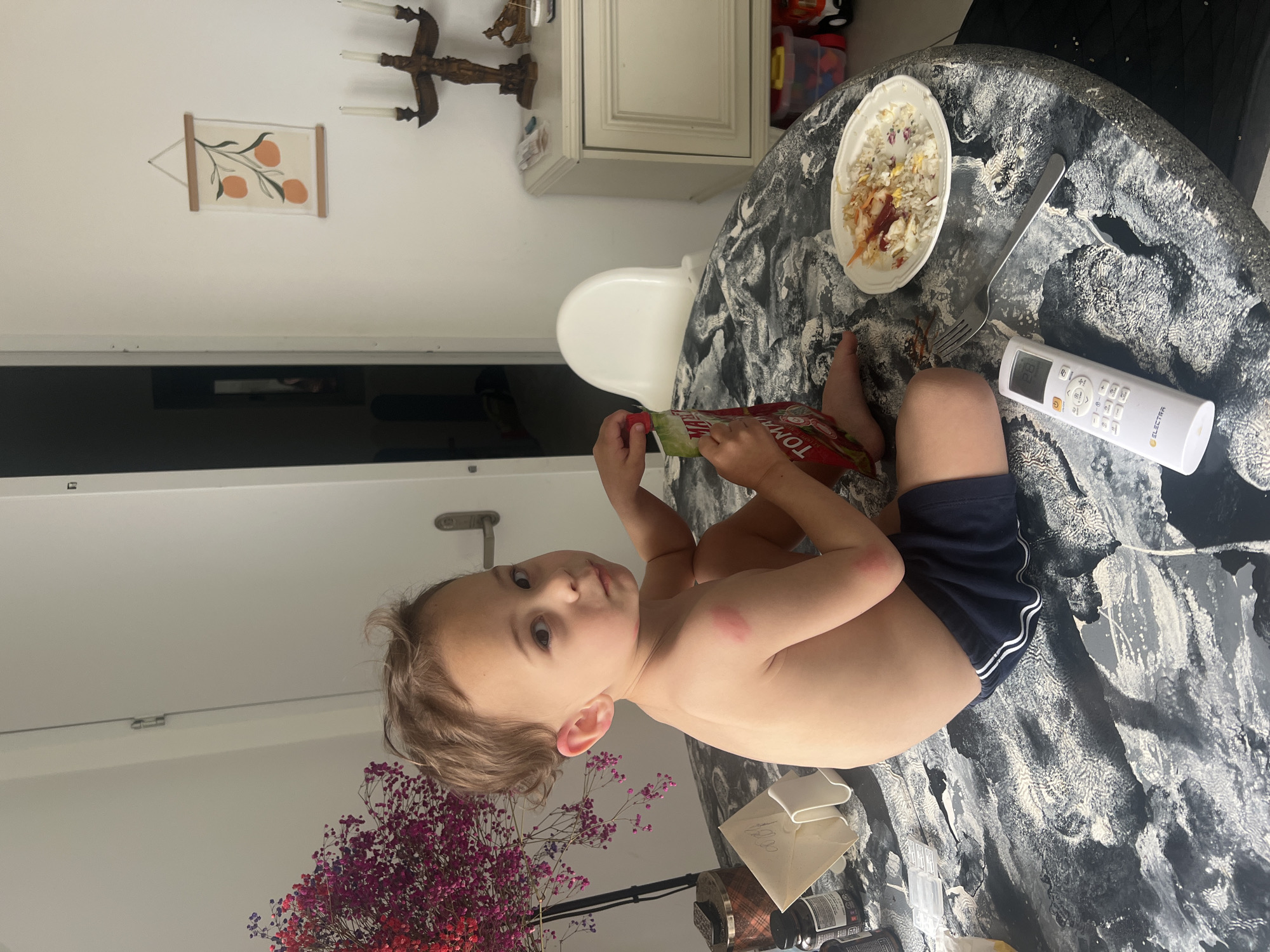 A young child sits at a black and white patterned table in what appears to be a home interior, possibly in Kiryat Bialik, Israel. The child is fair-skinned, shirtless, wearing dark blue shorts, and holding a packet of tomato sauce. He/She gazes towards the camera. The table also holds a half-eaten plate of food, a fork, and a white remote control. Further elements in the scene include a white chair, a white cabinet, wall art, and a decorative bouquet of red and purple flowers. The room has a white door and a light-colored wall.