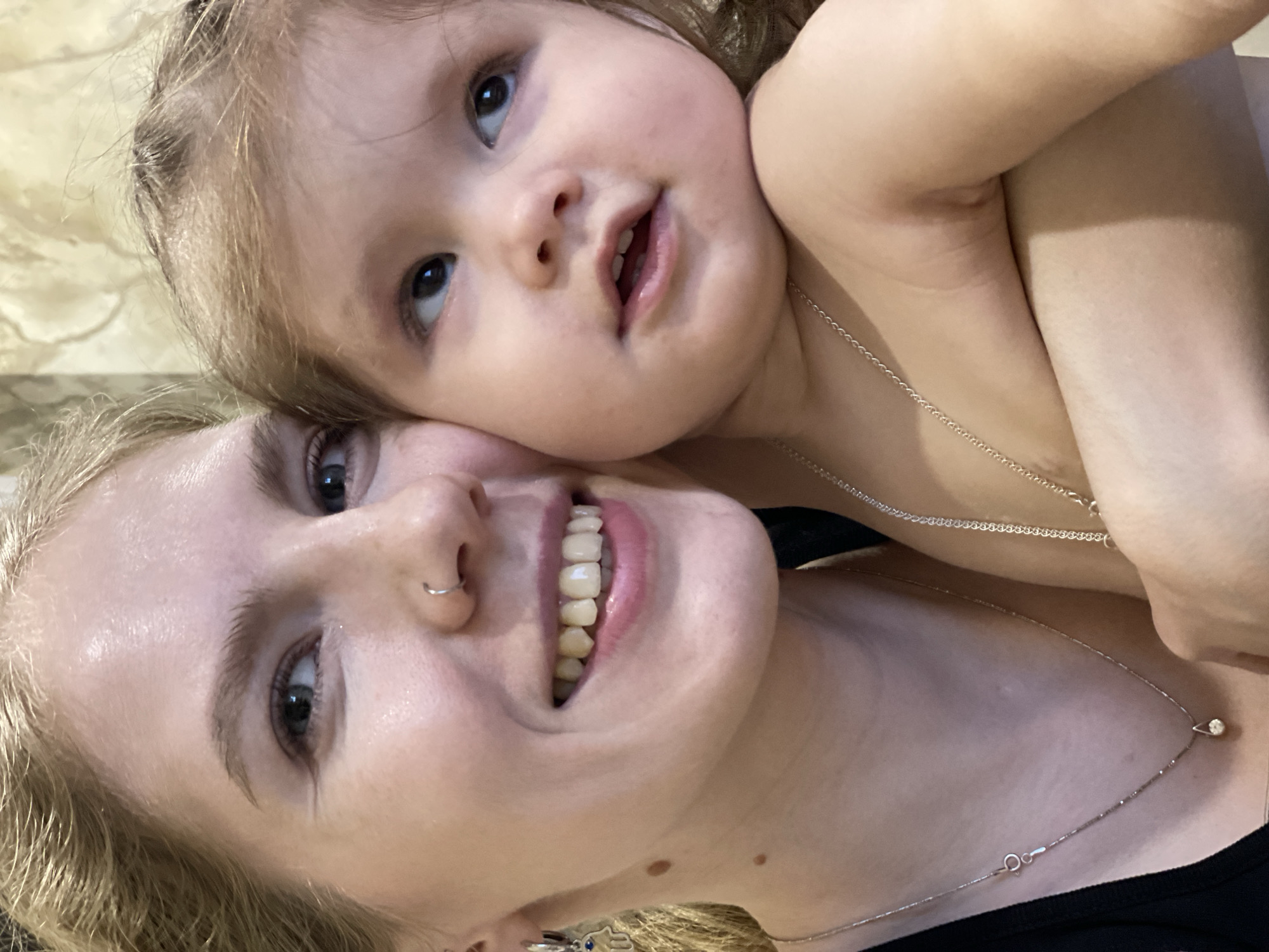 A close-up image depicts a woman and a young child, likely a mother and child, in an intimate embrace. The woman is smiling broadly, revealing her teeth and a nose ring. She is wearing a necklace. The child is nestled against her, also looking towards the camera with a slightly open mouth. The child is wearing a necklace. The image appears to be taken indoors with a light-colored background. There is no indication of the city Izhevsk, Russia.