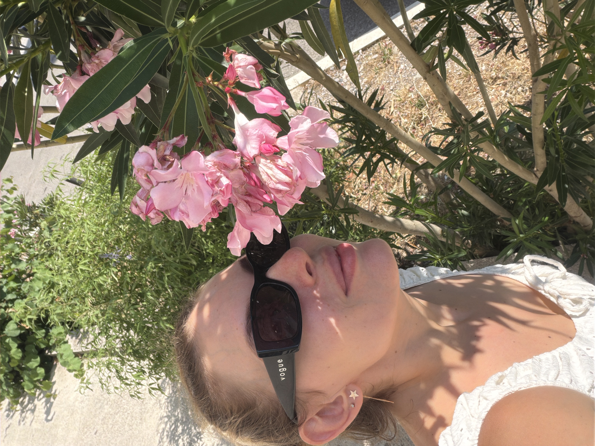 This media file features a close-up, overhead view of a young woman lying down outdoors, bathed in bright sunlight. She is wearing sunglasses with the brand name "Vogue" visible on the arm. Her head is tilted back, and her eyes are closed, suggesting relaxation or enjoyment of the sun. She has blonde hair and is wearing a white, textured top. An ear piercing with star-shaped studs is visible.
The scene is set amongst lush green foliage and pink oleander flowers, indicating a warm, possibly summer day. The ground appears to be covered in gravel and dry earth, typical of a Mediterranean environment. The lighting suggests it is daytime, with strong shadows cast by the surrounding plants, reinforcing the idea of bright sunshine. The overall impression is one of serene outdoor leisure in a Mediterranean location like Budva, Montenegro.