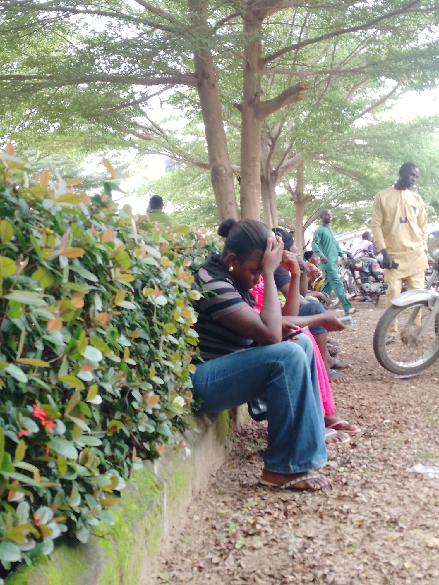 In Akwanga, Nigeria, this image captures a street scene under a canopy of trees. In the foreground, a woman in blue jeans and a black and white striped shirt sits with her hand on her forehead, holding a phone. To her right, another person in a pink garment is seated. Further back, a man in a yellow traditional outfit stands near a motorcycle. Another man in a green outfit walks in the background. The ground is covered with dirt and fallen leaves, with a moss-covered concrete barrier running along the left side of the image. Lush green foliage borders the barrier.