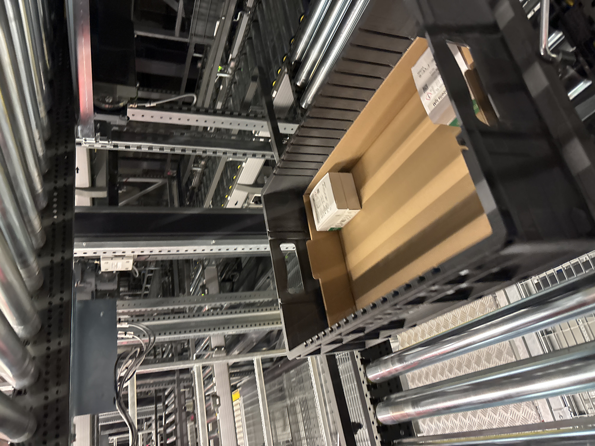 The image depicts an automated warehouse system. In the foreground, a dark plastic bin is situated on a conveyor belt system composed of metal rollers. The bin contains a small cardboard box. In the background, a complex network of shelving units, conveyor belts, and machinery fills the space, indicative of a large-scale logistics operation. The environment appears to be an industrial indoor setting. The image does not provide specific visual cues to determine its location in Mannheim, Germany.