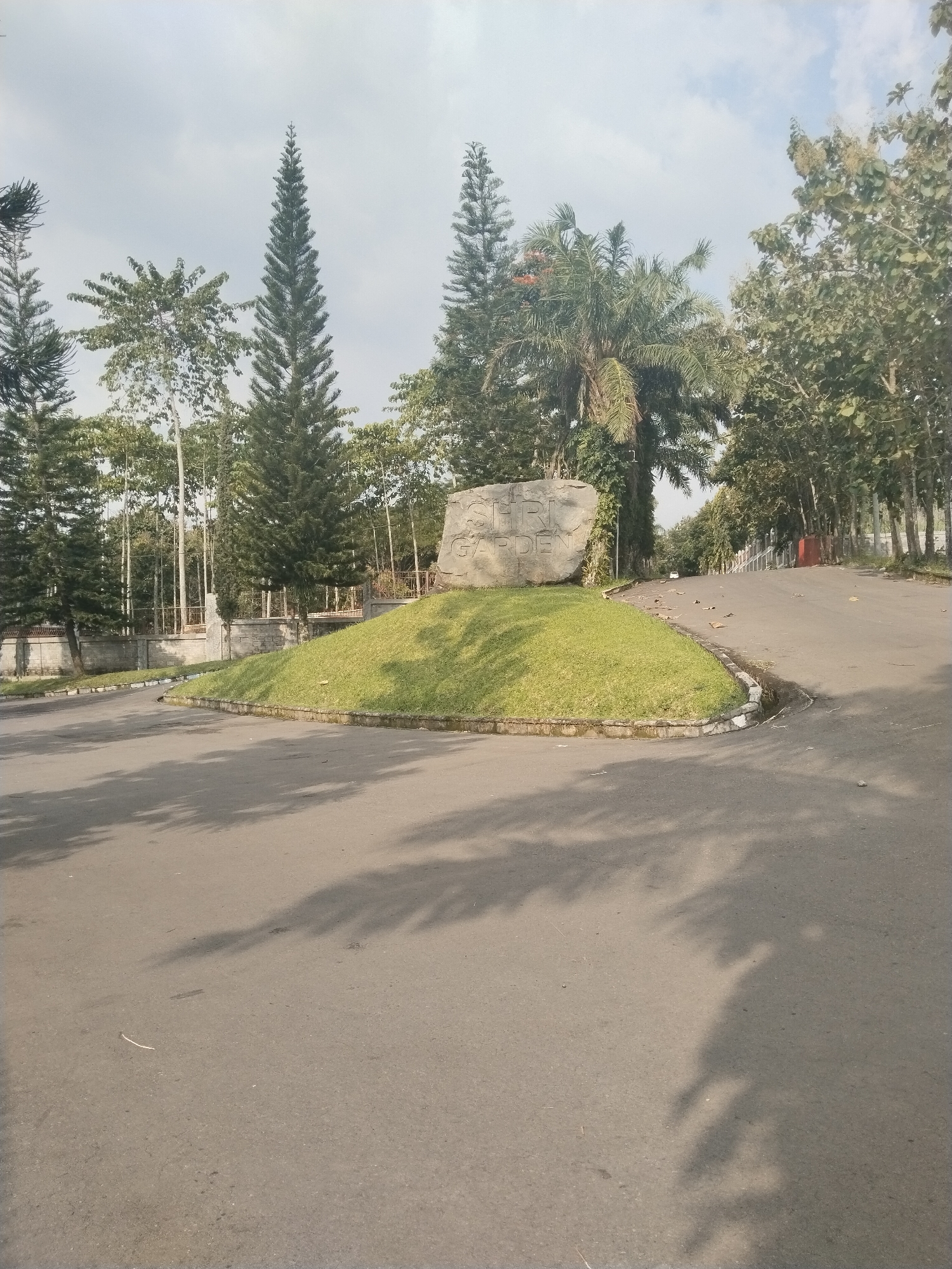 The image captures an outdoor scene at what appears to be an entrance or a fork in a road within a landscaped area, set in Tohkuning, Indonesia. In the center of the frame is a rounded, grassy mound, which serves as a median for the asphalt road. Prominently placed on this mound is a large, light-colored stone with text carved into its surface, reading "SHRI GARDEN."
The scene is rich with various types of trees. Tall, slender pine-like trees (conifers) are visible on both sides of the road and in the background, contrasting with broader-leaved deciduous trees and a large palm tree situated behind the stone sign. The sky above is bright with scattered clouds, indicating a clear day. Distinct shadows of trees are cast across the asphalt road in the foreground, suggesting it is either morning or early afternoon.
On the left side of the image, behind the road and grassy verge, there is a low stone wall topped with a metal fence, partially hidden by the foliage. The road to the right of the grassy mound slopes gently uphill and curves out of view, lined by more trees, some with tall, slender trunks. Scattered leaves are visible on this section of the road. No people or specific activities are depicted in the static landscape. The overall impression is one of a well-maintained and green environment, likely a park, residential estate, or resort.