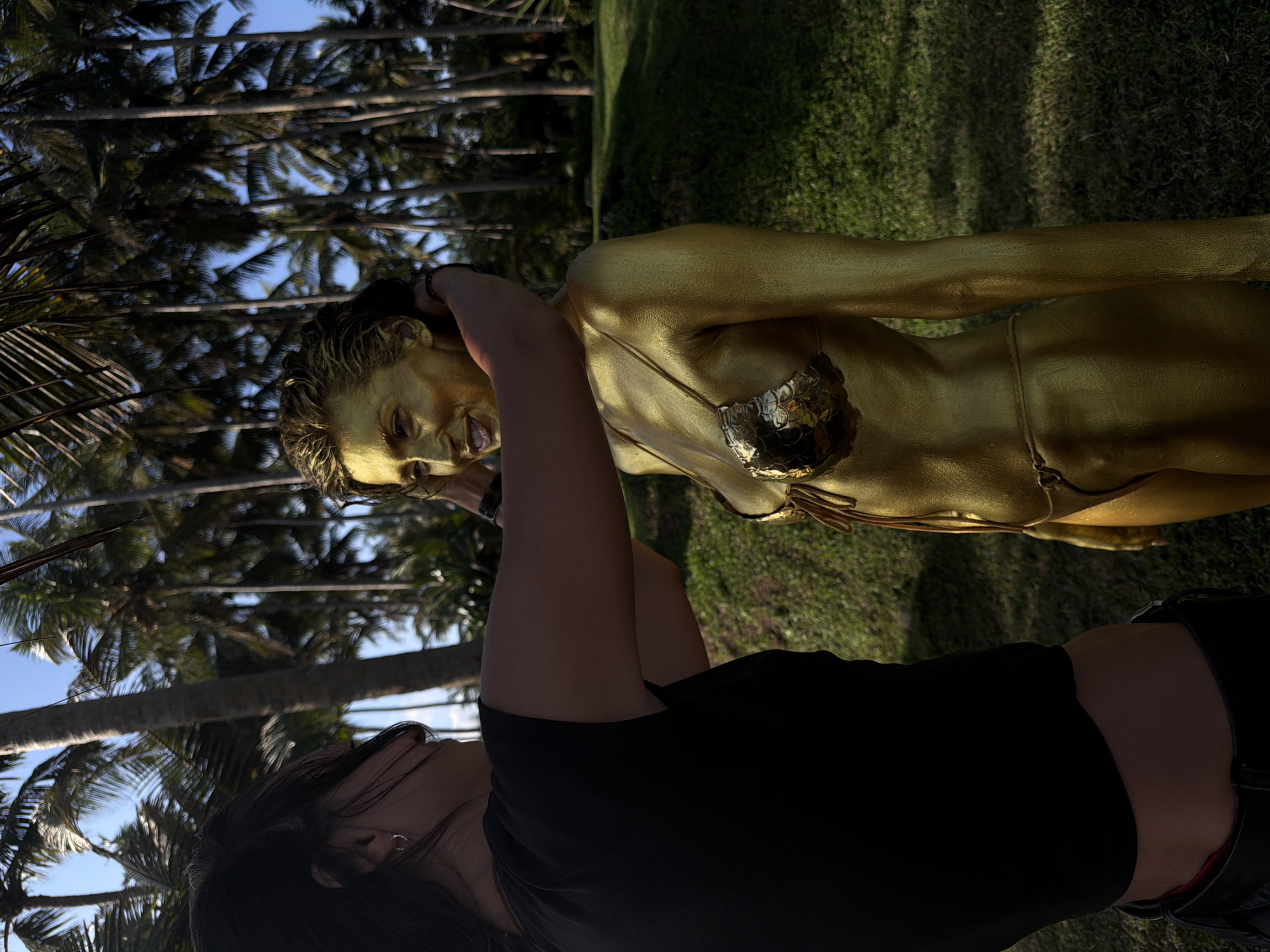 A person in a black shirt and dark hair is angled toward the camera and is holding a camera aimed at a person painted gold, wearing a gold bikini made of shiny plates, and posing. Behind the person in gold are palm trees and a patch of green grass. The location is Kerambitan, Indonesia.