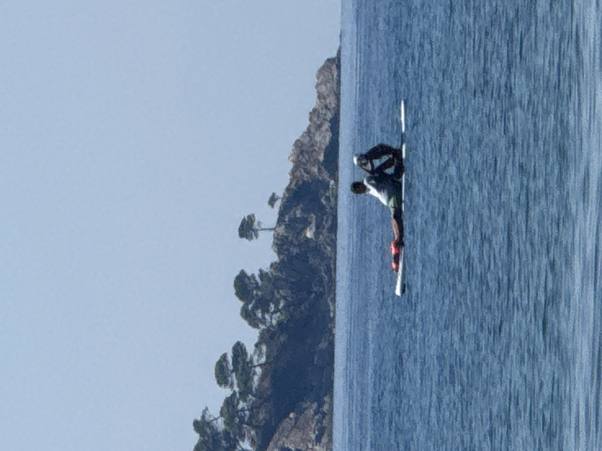 In the city of Buljarica, Montenegro, two individuals are positioned on a stand-up paddleboard in the calm, blue sea. The person in front is seated and appears to be wearing a white top. The person behind is also seated, wearing a dark top and a white helmet. Both individuals are facing to the left. A red life vest is visible on the paddleboard. In the background, a rocky coastline covered with green trees stretches across the horizon. The sky is a clear, pale blue.