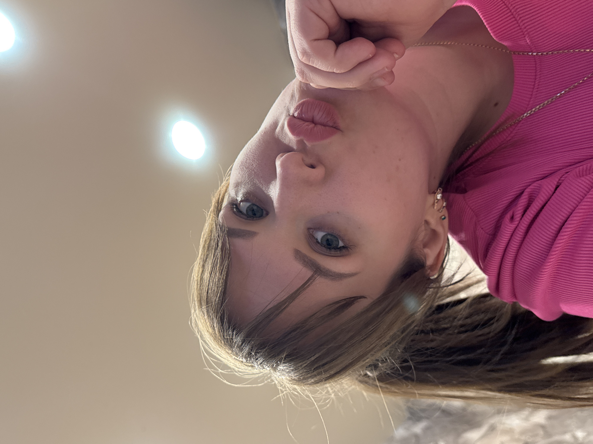 This media file presents an upside-down, close-up portrait of a person, likely a young woman, captured indoors. The subject has light brown hair that falls around her face and over her shoulders, fair skin, and light blue or green eyes that are open and directly engaging with the camera. Her lips are puckered in a "kissing" or "duck face" expression, conveying a playful or affectionate mood.
She is dressed in a bright pink ribbed top or sweater, and a delicate gold chain necklace is visible around her neck. Small, stud-style earrings can be seen in her ears. Her left hand is partially visible near her chin, with fingers slightly curled.
The background is a plain, light-colored ceiling, possibly beige or off-white, which is brightly illuminated by two prominent, circular recessed light fixtures. The lighting is artificial and bright. The inverted orientation of the image suggests either the person was looking up at the camera while taking the selfie, or the image has been rotated post-capture. The setting is clearly an interior space, with no external environmental cues. No text is visible in the image. The location context provided is Chaikovsky, Russia.