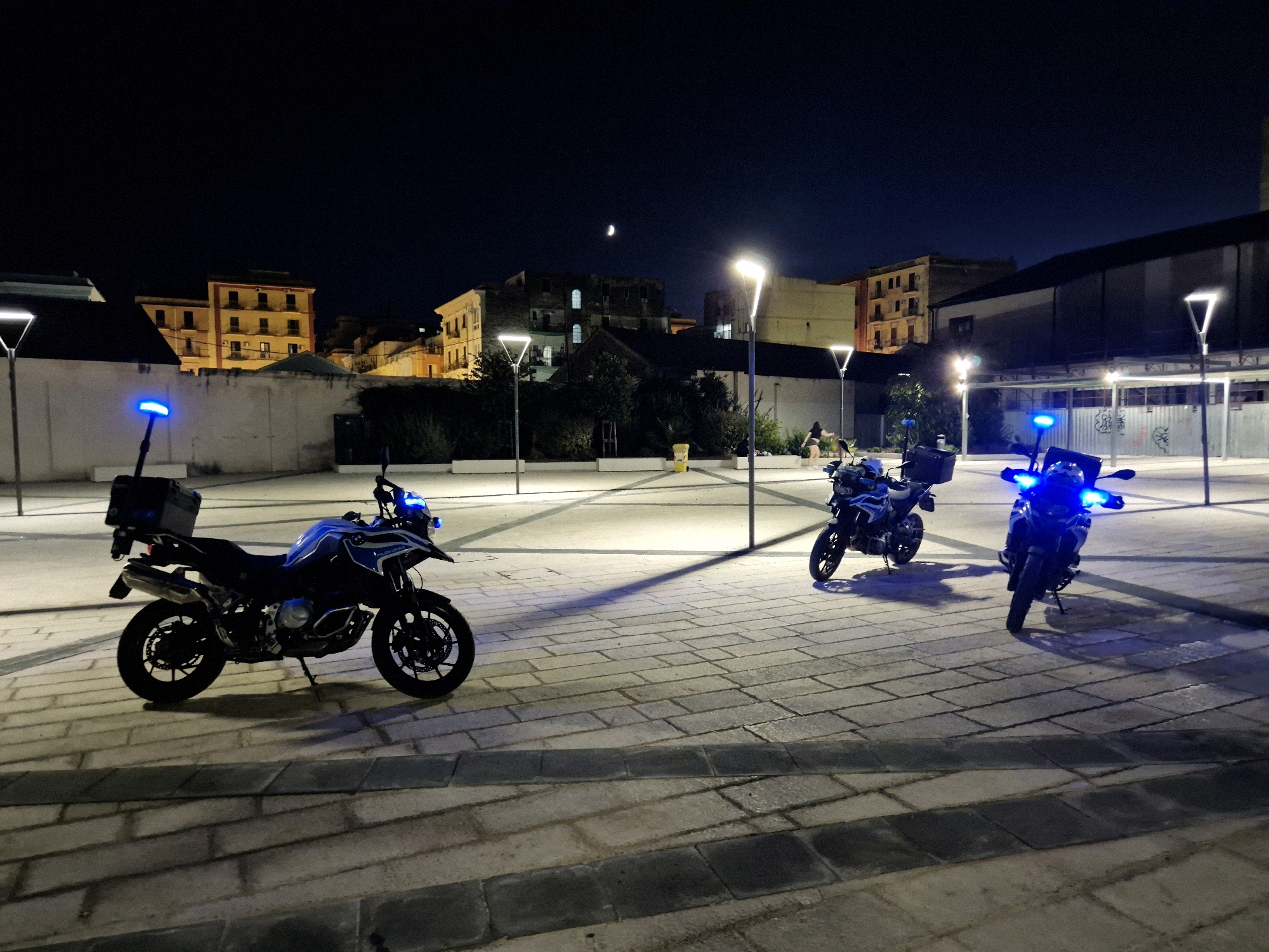 At night, three police motorcycles with blue flashing lights are parked in a paved square in Taranto, Italy. The motorcycles are positioned on the right side of the image, with the one closest to the viewer on the left. In the background, buildings with illuminated windows and streetlights can be seen. The sky is dark, with a crescent moon visible. The square appears to be empty of people, except for two figures walking in the distance on the right side of the image.
