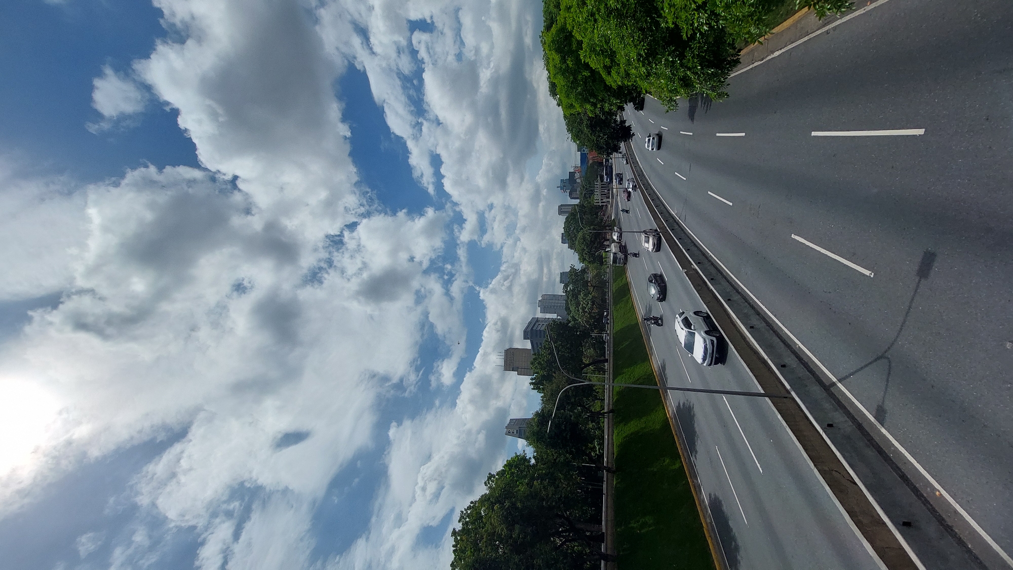 The image captures a view of the Francisco Fajardo Highway in Caracas, Venezuela, oriented to display the sky on the left and the road running vertically on the right. The sky is filled with a mix of blue and white clouds. The highway has multiple lanes with cars and motorcycles moving along it. Buildings can be seen between the highway and the sky. Trees are present on either side of the highway, creating a green divide. The sun can be seen shining in the bottom-left corner of the image.