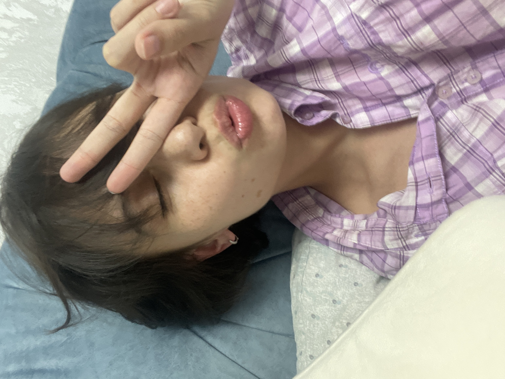 A high-angle, close-up shot depicts a person lying down in what appears to be an indoor setting, such as a bed or sofa, in Izhevsk, Russia. The individual, whose gender is not explicitly discernible but appears youthful, has dark hair and prominent freckles scattered across their nose and cheeks. They are wearing a purple and white plaid (checkered) button-up shirt.
The person's right hand is raised, forming a "peace sign" (V-sign) with their fingers, which are partially obscuring their eyes. Their lips are glossy, slightly parted, and pursed into a pout, suggesting a playful or "kiss face" expression. A small, silver-colored hoop earring is visible in their left ear. The head rests on a blue textured fabric, likely a pillow or blanket.
The background features several soft textiles, including the blue fabric, a pale, subtly patterned fabric visible beneath the shirt, and a plain, light-colored (possibly white or cream) soft surface to the person's right. The lighting is soft and diffused, characteristic of indoor ambient light. The overall mood conveyed by the pose and expression is relaxed, playful, or perhaps a little shy. No visible text is present in the image.