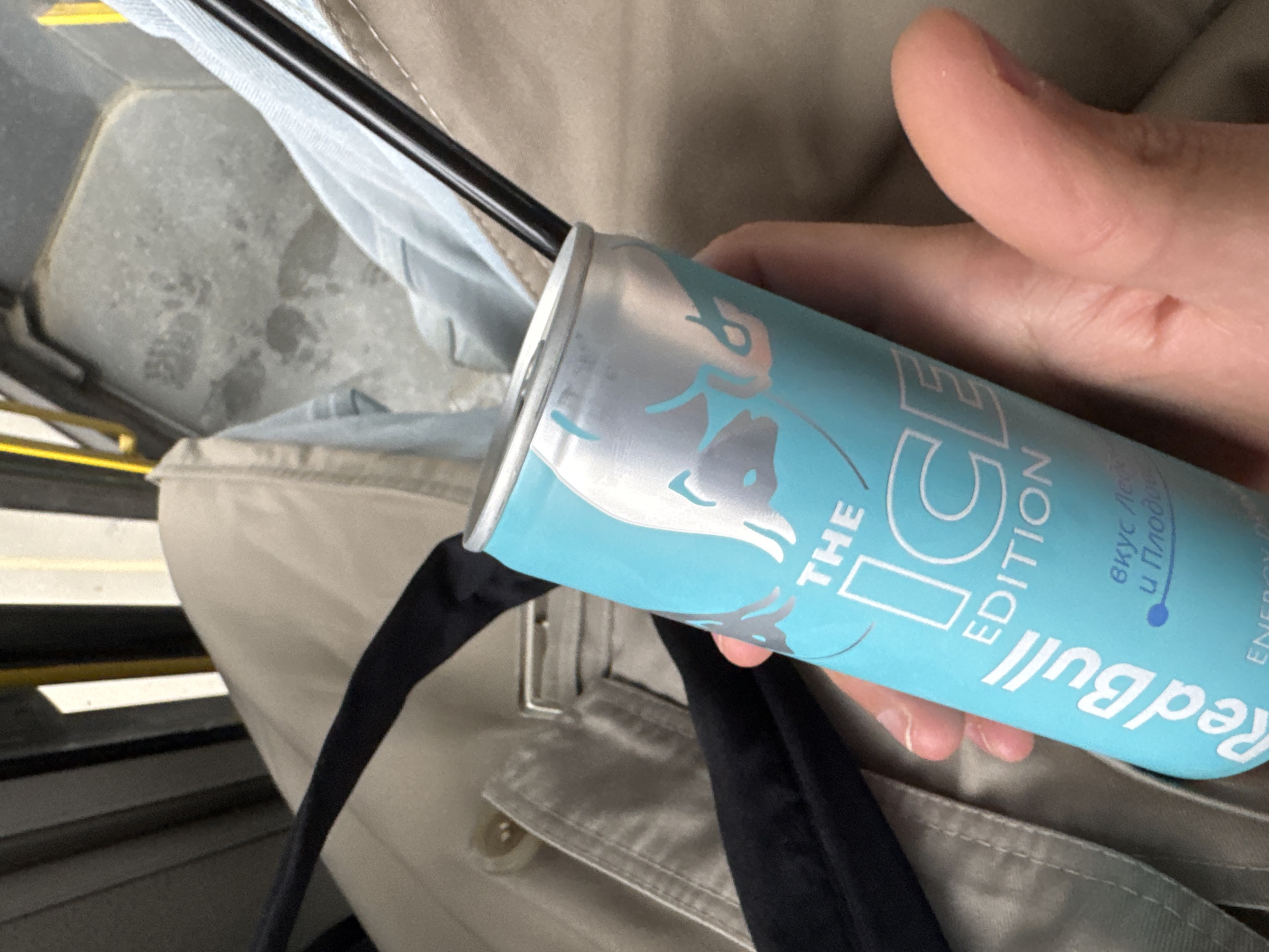 A hand is holding a light blue can of Red Bull "The Ice Edition" with a black straw inserted. The can is tilted. The background suggests a public transport setting, possibly a bus or tram, with a beige seat and a metal bar visible. The text on the can includes Russian Cyrillic characters, indicating it may have been purchased in a region where Russian is spoken. The location might be Sofia, Bulgaria, but it is difficult to determine from the picture alone. The focus is on the can of Red Bull held by the hand.