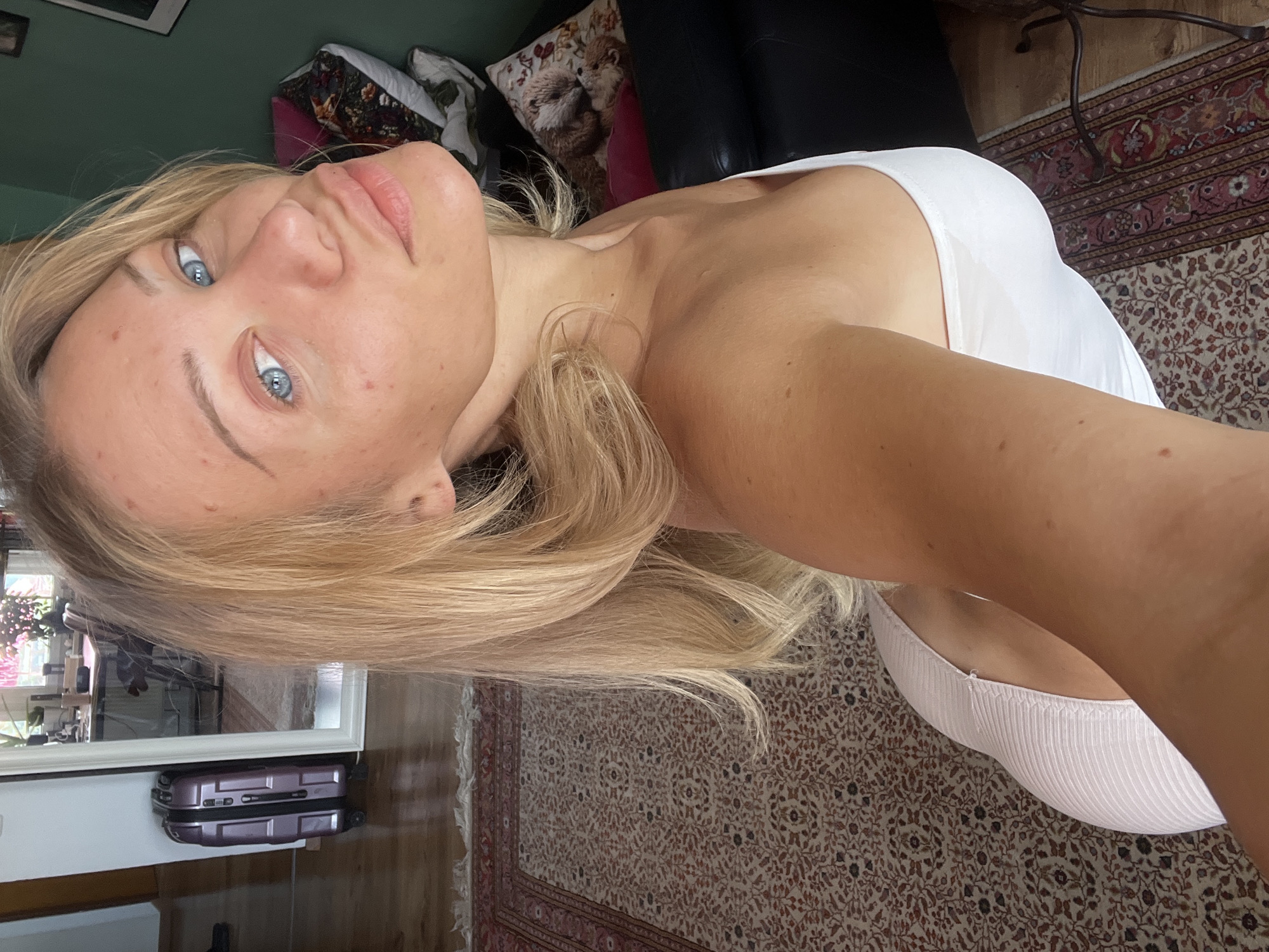 A woman with blonde hair and blue eyes is taking a selfie in a room that appears to be a living room. She is wearing a white strapless top. Behind her, there is a suitcase, a large mirror, a patterned rug, and a dark green wall with pillows on a sofa. The scene is lit by natural light. The location is in Mühldorf a. Inn, Germany.