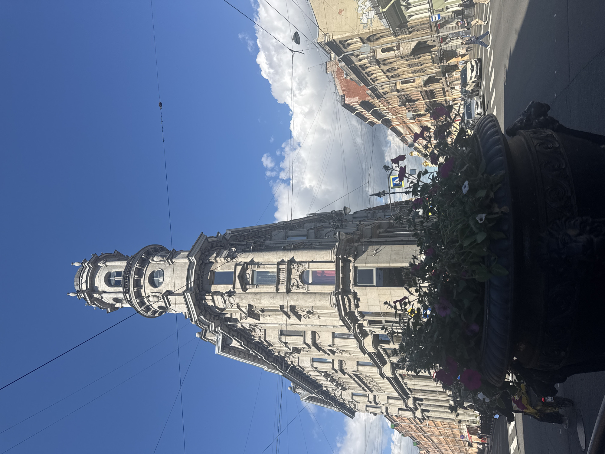 The image depicts a street view in Saint Petersburg, Russia, featuring a prominent, ornate, triangular building with a domed tower. The building's facade is adorned with classical architectural details, including columns, decorative carvings, and numerous windows. In the foreground, a large decorative planter filled with flowers partially obscures the lower portion of the building and the street.
The street is paved and shows pedestrian crossings. Several cars are visible on the street, including a white sedan and a white SUV. Pedestrians are crossing the street at a designated crosswalk. Along the right side of the street, a row of similarly styled buildings stretches into the distance. The sky is a bright blue with scattered white clouds. Overhead, several dark electrical or utility wires crisscross the scene. The lighting suggests it is daytime, with shadows cast on the street.