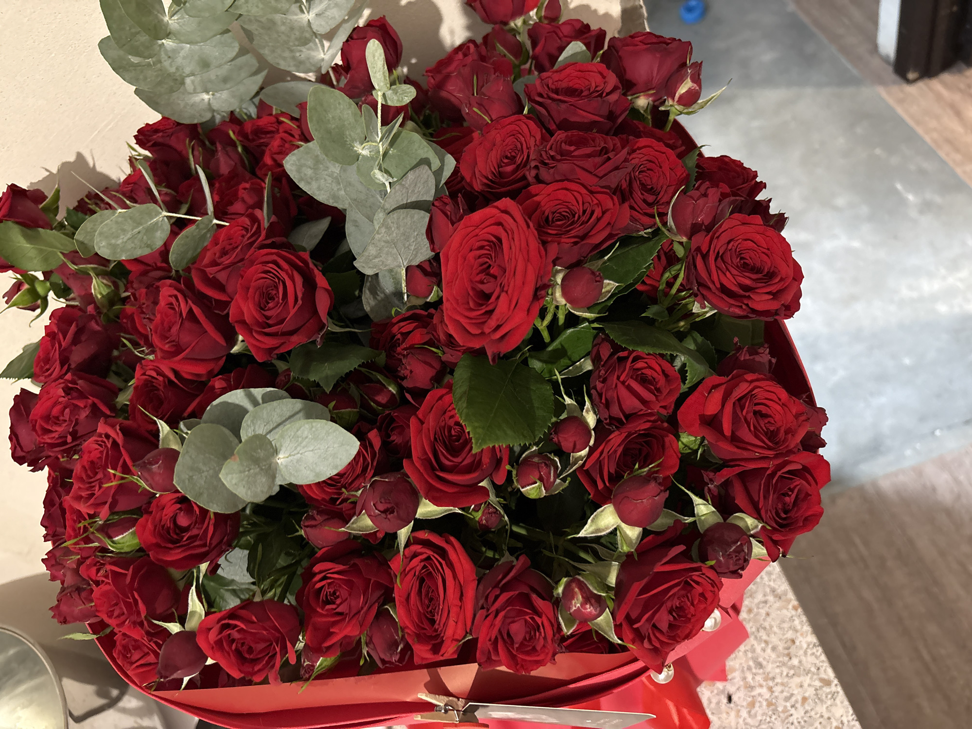 The image shows a close-up, high-angle view of a bouquet of red roses, arranged in a red box. The roses are a deep red color and some are fully bloomed while others are still buds. There are also several sprigs of grey-green eucalyptus leaves interspersed among the roses. The bouquet is tied with a red ribbon and a small white tag is attached to it. The background is a light-colored wall. The foreground shows a speckled surface that appears to be a countertop or table, and a hint of a light blue object in the bottom right corner. The scene does not provide any information that would allow for the identification of the city of Rostov-on-Don, Russia.
