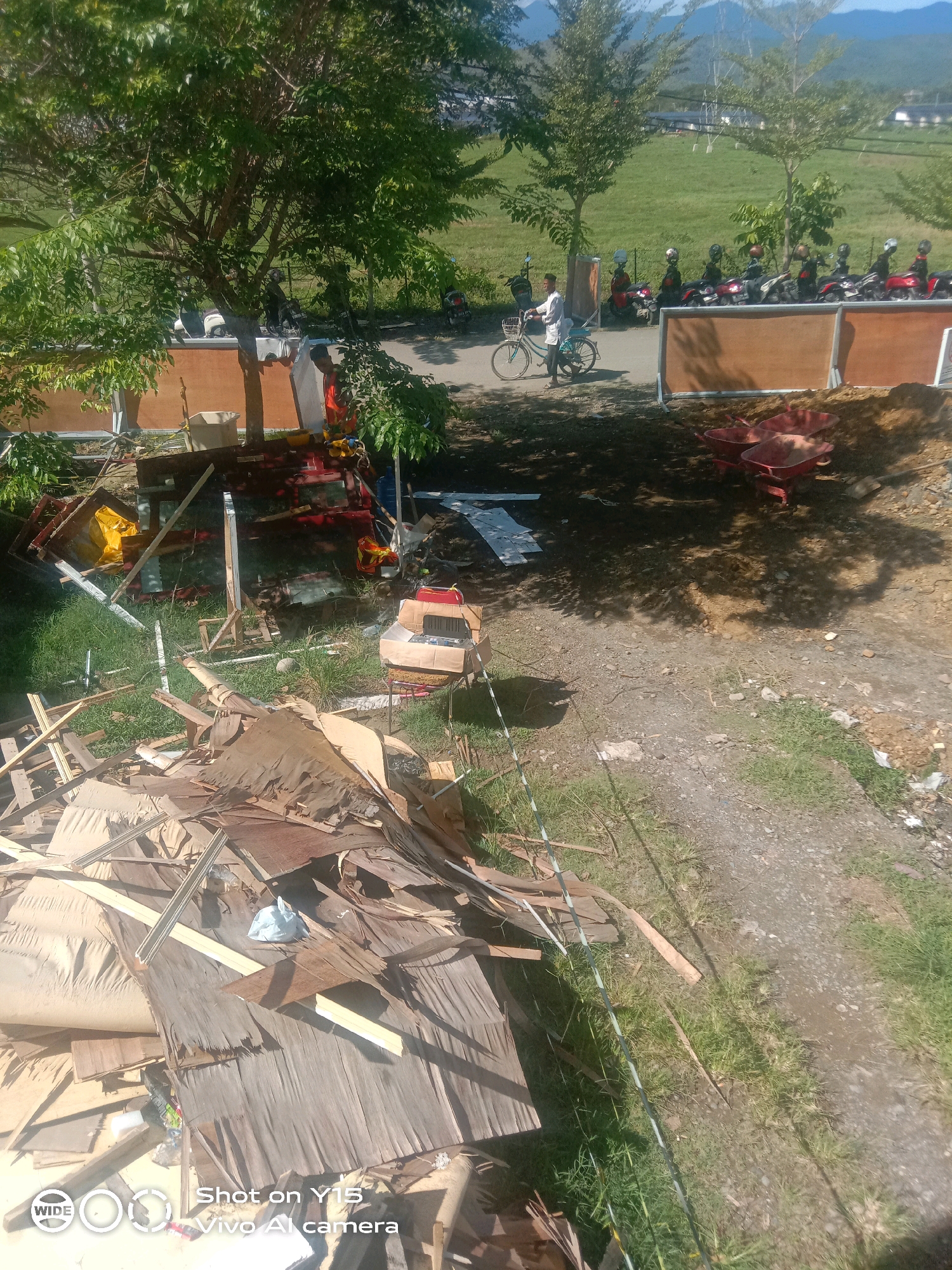 The image shows a construction or demolition site in the city of Meureudu, Indonesia. In the foreground, there is a large pile of debris, primarily consisting of wood and what appears to be broken furniture or building materials. A white plastic bag is visible on top of the debris. Behind the debris, a dirt path leads into the scene.
Further into the image, several red wheelbarrows are present, suggesting ongoing work or materials being moved. A group of motorcycles are parked in a line, some with helmets on. A person wearing a light-colored shirt and dark pants is walking a bicycle. In the background, trees and a grassy field are visible, with some buildings and a mountainous landscape in the distance. The scene is illuminated by sunlight, casting shadows on the ground. The overall impression is one of a busy outdoor work area with signs of construction or cleanup activities.