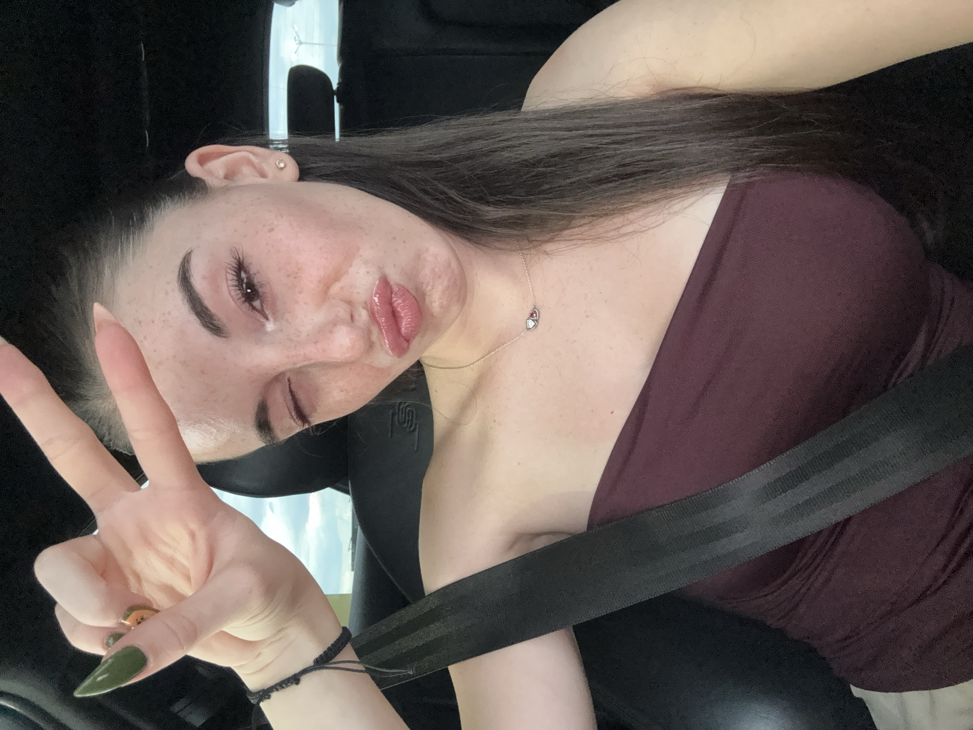 A woman is seen from the chest up, inside a vehicle, with a seatbelt across her. She is making a peace sign with her right hand, and winking with her left eye. She has on a strapless burgundy top and a necklace with two heart-shaped pendants. Her hair is dark brown and pulled back. Her skin is fair with freckles. The background appears to be the interior of a car, with a dark headrest visible. The sky is visible through the car window, suggesting it is daytime. The image has been cropped to focus on the woman.