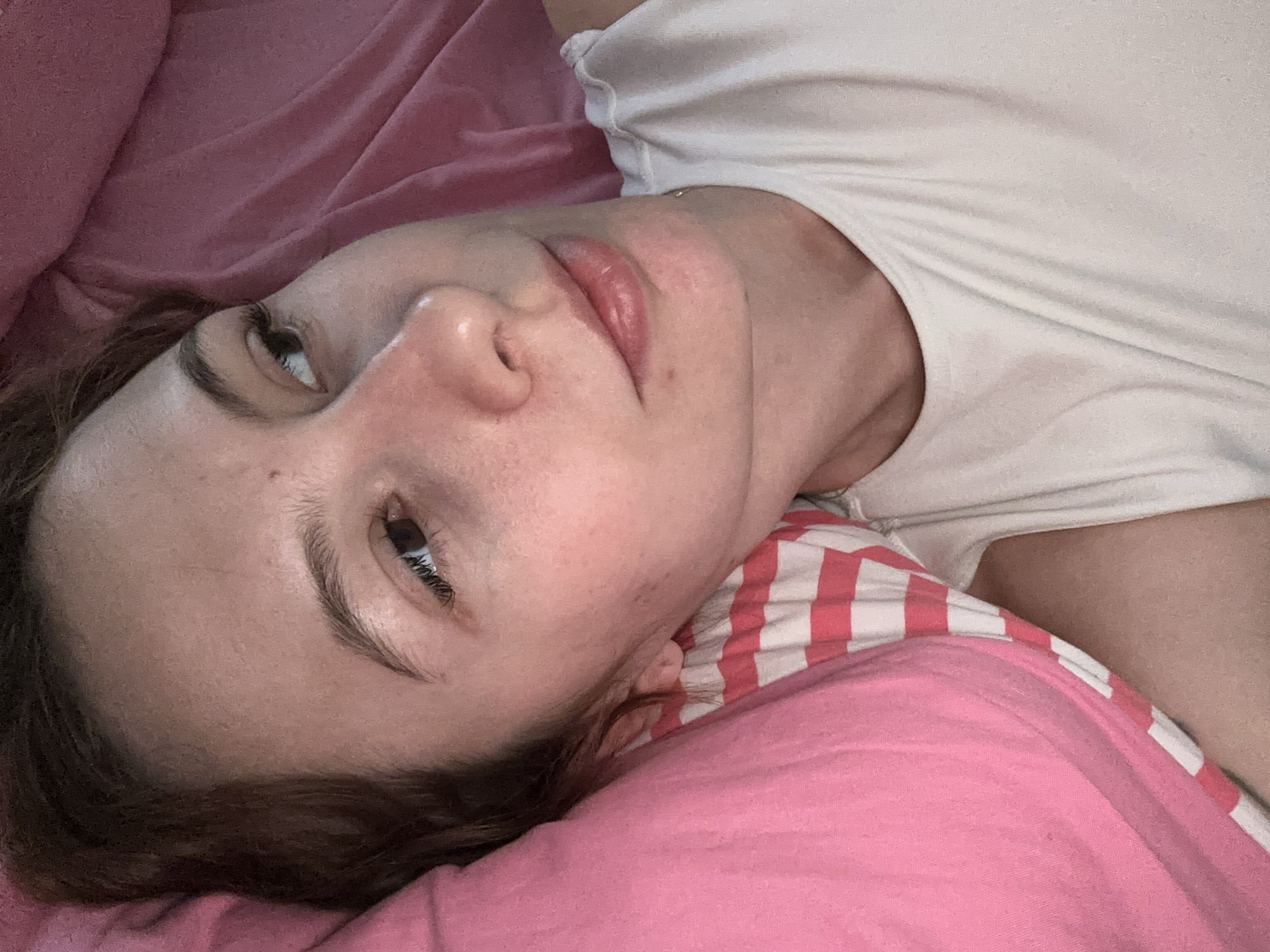 A close-up, high-angle shot captures a person, likely a woman, lying down indoors, seemingly in a bed. Her head rests on a plain pink pillow, while a second pillow with pink and white stripes is visible behind her neck. She wears a simple, light-colored (possibly white or off-white) t-shirt.
Her face is the central focus, turned slightly to the viewer's left, with her eyes gazing upwards and to the side, past the camera. Her dark brown hair is spread around her head against the pink bedding. Her lips are slightly parted, revealing their natural rosy hue. Her facial expression appears neutral and relaxed, suggesting a state of rest or quiet thought. Natural skin textures and minor blemishes are visible.
The lighting in the image is soft and diffused, indicating an indoor setting, likely a bedroom. There are no harsh shadows, suggesting either ambient room lighting or an overcast daylight scenario. The overall mood is calm and intimate. No specific textual elements or strong environmental cues, such as weather conditions or distinct architectural features, are discernible to pinpoint the exact time of day or location beyond a generic indoor setting.