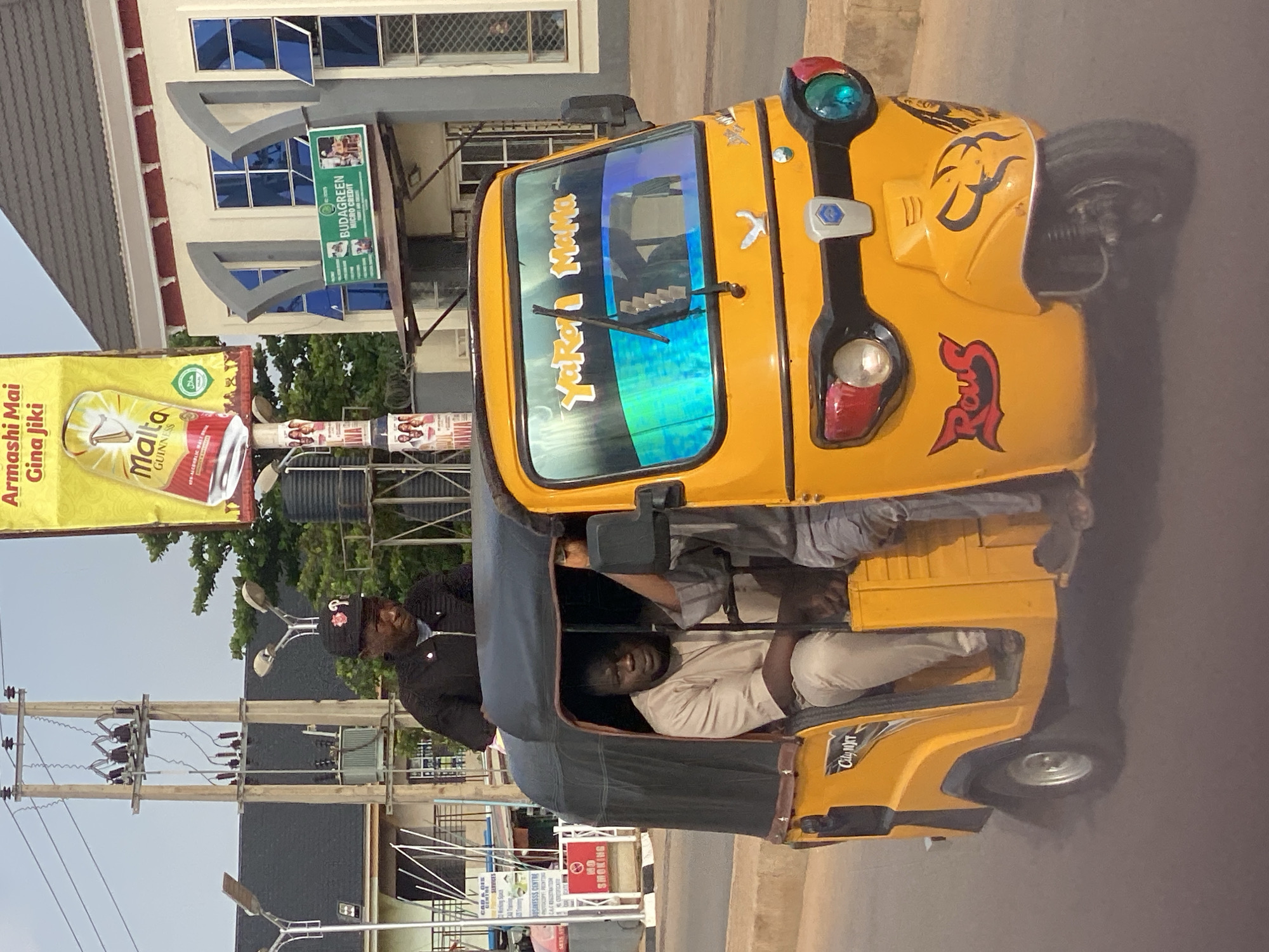 In the city of Sabon Tasha, Nigeria, a bright yellow auto-rickshaw, also known as a Keke Napep, is the central focus of the image. The Keke Napep is in motion, indicated by the slightly blurred background and the tire kicking up a dust cloud.
There are three individuals visible in and around the vehicle. The driver, a man wearing a black jacket and a black baseball cap with red floral embroidery and a white emblem, is seated at the front. He appears to be looking forward. Inside the passenger compartment, a man in a light-colored, loose-fitting shirt and pants is seated on the left. To his right, another person, partially obscured, is also seated.
The Keke Napep has several distinctive features. The windshield is decorated with a reflective film that displays a gradient of blue, green, and yellow. White lettering in a stylized font reads "Yaro Mama" on the upper part of the windshield. The front of the vehicle has a silver emblem and two headlights, one red and one blue. A black and red "Rous" logo is visible on the side, near the front wheel. The body of the Keke Napep is adorned with a tribal or animalistic graphic design in black on the front fender. The word "City" and possibly "TKT" are visible in white lettering on the side of the passenger compartment.
The background shows elements of an urban environment. A tall utility pole with multiple transformers and electrical wires is prominent on the left side of the image. In the distance, a building with a dark roof and light-colored walls features blue-tinted windows. A large yellow billboard with text in what appears to be a local language and an image of a can of Malta Guinness is also visible. Signage for "BUDAGREEN MICRO CREDIT" is affixed to the building. Trees and other vegetation are present in the background. The overall scene suggests a typical street view in a Nigerian city.