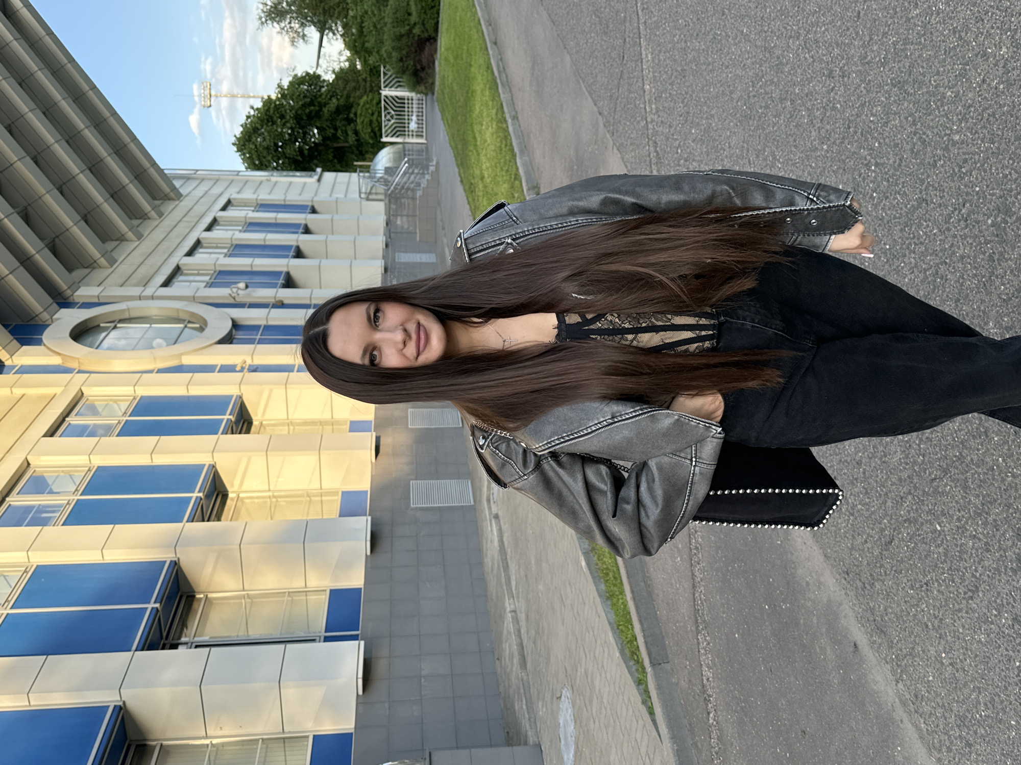 A young woman with long, dark brown hair is pictured standing outdoors, smiling faintly towards the camera. She is dressed in a dark grey, distressed-look leather jacket adorned with studs or decorative elements on the collar and cuffs. Underneath, she wears a black lace or mesh top, and dark, possibly black, jeans or trousers. A thin silver chain with a cross pendant is visible around her neck. She carries a black handbag with silver stud detailing along its edge. Her fingernails are painted a light, neutral color.
The background on her left features a modern building with a striking facade composed of grey and white panels, interspersed with prominent blue-tinted windows, some rectangular and one circular. The building's design is angular and contemporary. To her right, a paved walkway or asphalt surface stretches back, bordered by a strip of green grass and several trees. A white metal fence or gate is partially visible in the mid-ground, and a tall stadium-style light pole can be seen in the distance against a light blue sky with scattered clouds. The scene appears to be set during daytime, possibly late afternoon, with soft, natural light illuminating the surroundings. There is no visible text in the image.