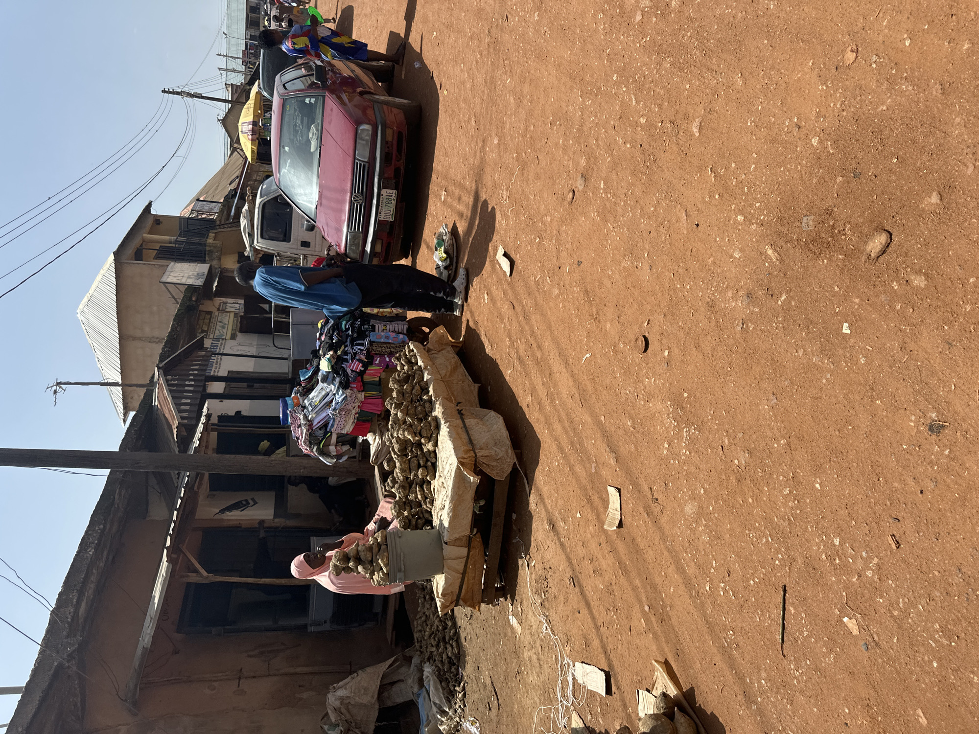 In Bukuru, Nigeria, a busy outdoor market scene unfolds on a dirt road under a bright, clear sky. In the foreground, a stall displays a large quantity of root vegetables, some piled high in a grey bucket and others neatly arranged on a table covered with a light-colored cloth. A woman wearing a pink headscarf is seated behind the stall, facing left and appearing to be engaged in the market activities. To the right of the stall, a red Volkswagen sedan is parked, its front bumper facing the viewer. A man in a blue shirt and dark pants stands near the car, his attention directed downwards. Further to the right, a woman in a vibrantly patterned dress walks away from the camera, holding a green object in her right hand. In the background, a row of market stalls and buildings line the street, with various goods displayed for sale. A white van is parked behind the red sedan, and several other vehicles and people are visible further down the road, suggesting a bustling commercial area. The ground is a dusty, reddish-brown dirt, scattered with a few pieces of debris. The overall impression is one of a vibrant, everyday market in a Nigerian city.