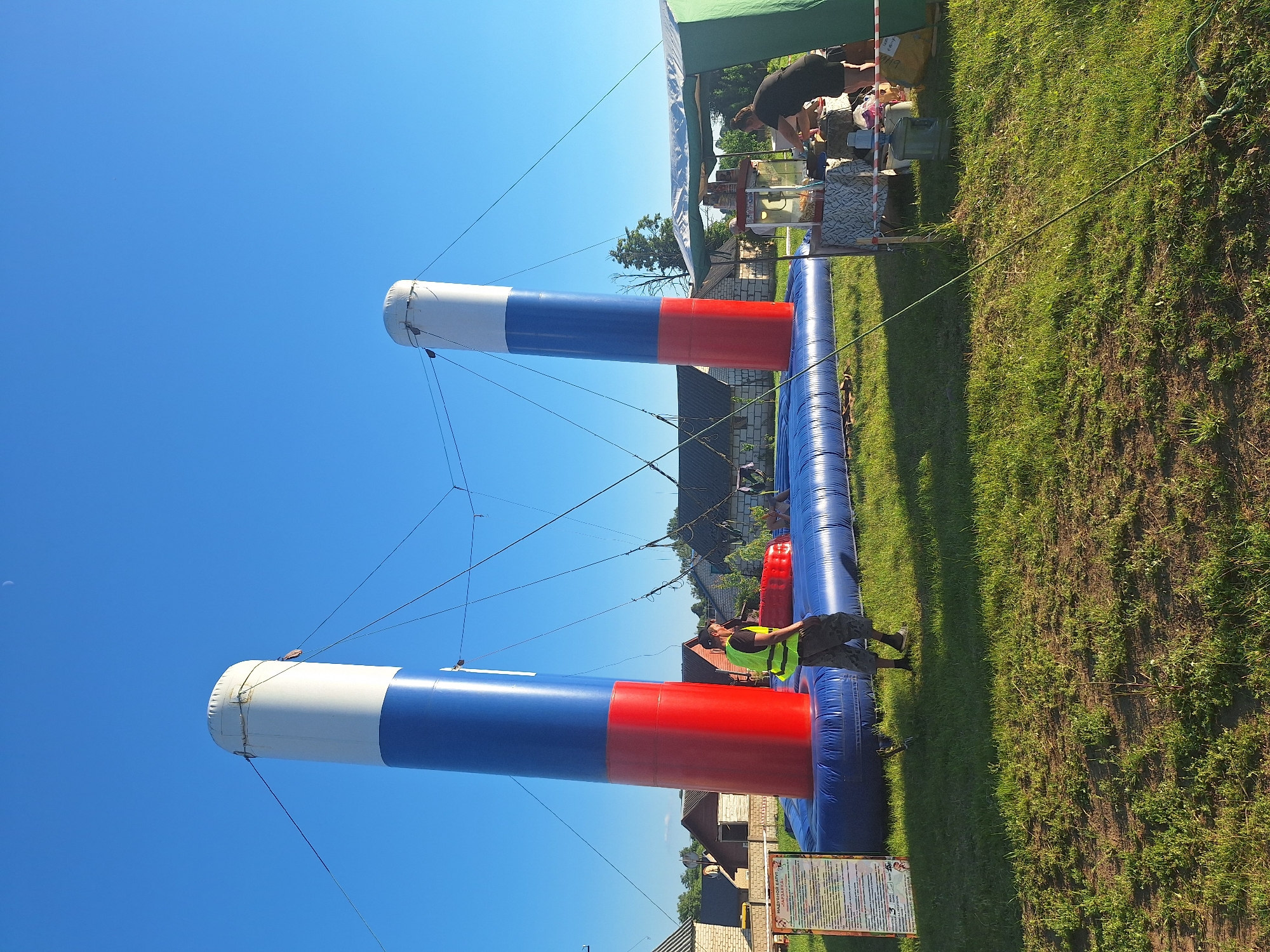 This image captures an outdoor event in Цімкавіцкі сельскі Савет, Belarus, on a clear day with a bright blue sky and a sliver of a moon visible. In the foreground, a large inflatable attraction, resembling two tall columns decorated with the colors of the Russian flag (red, white, and blue), is set up on a blue inflatable base. A man wearing a high-visibility yellow vest and camouflage pants stands near the inflatable structure, observing the scene. To the right of the attraction, a temporary canopy tent is erected, beneath which a woman in a black t-shirt and shorts is attending to a food stall. Several people are visible around the food stall, some appearing to be customers. The ground is covered in green grass, with some patches of exposed soil. In the background, several houses with brick and wooden exteriors are visible, suggesting a rural or semi-rural setting. The overall atmosphere appears to be a lively outdoor gathering or festival.