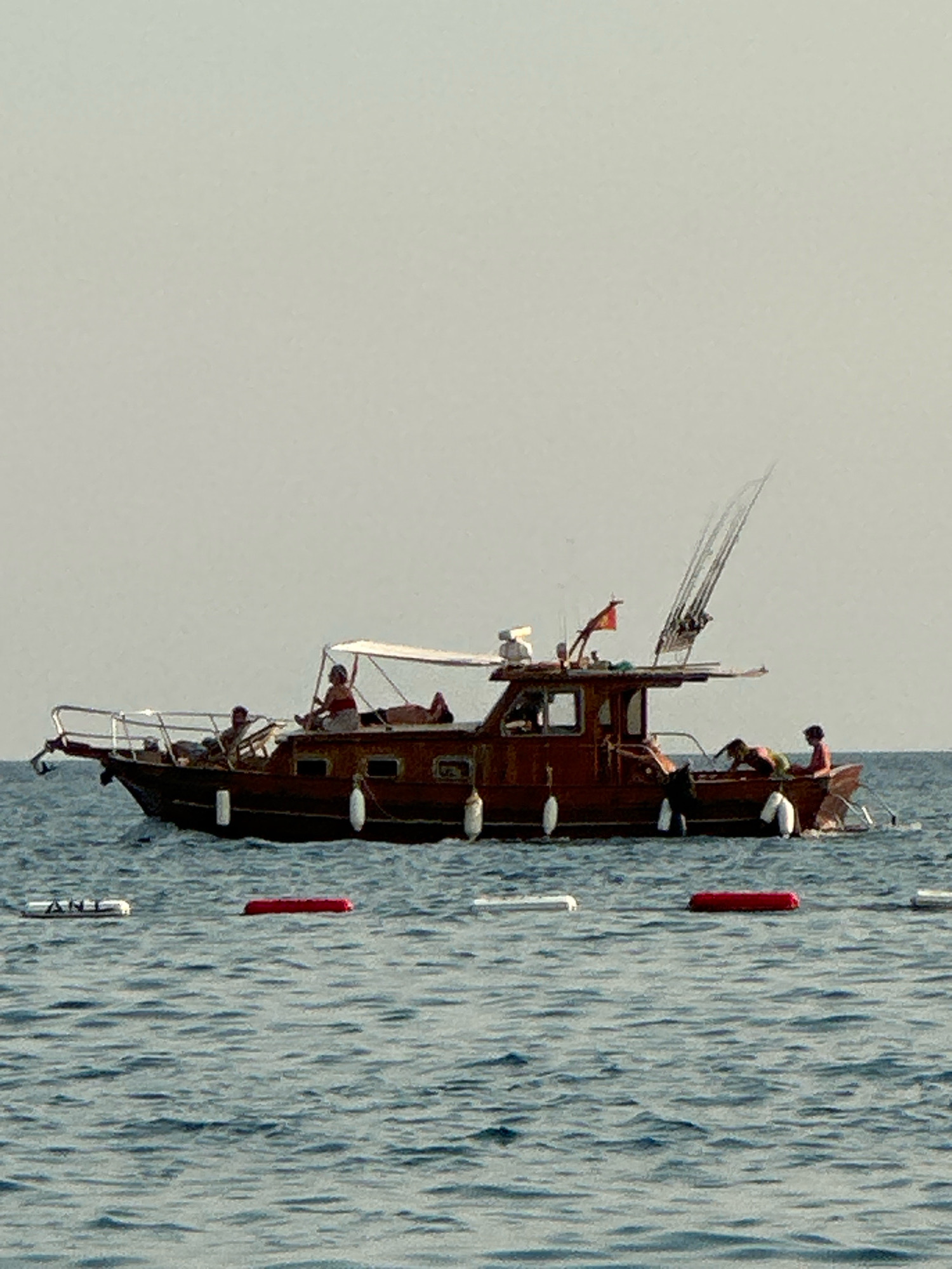 A medium shot captures a wooden boat in the water near Boreti, Montenegro. The boat, which appears to be a fishing vessel, has several people on board. On the foredeck, two individuals are visible, one sitting and the other lying down. Towards the stern, a person is seated under a canopy, and another is leaning over the side. In the rear of the boat, two more individuals are present, one standing and another sitting. The boat is equipped with fishing rods and a flag. In the foreground, several buoys are floating in the water, some red and some white. The water is calm and reflects the overcast sky. The overall scene suggests a leisurely day out on the water.