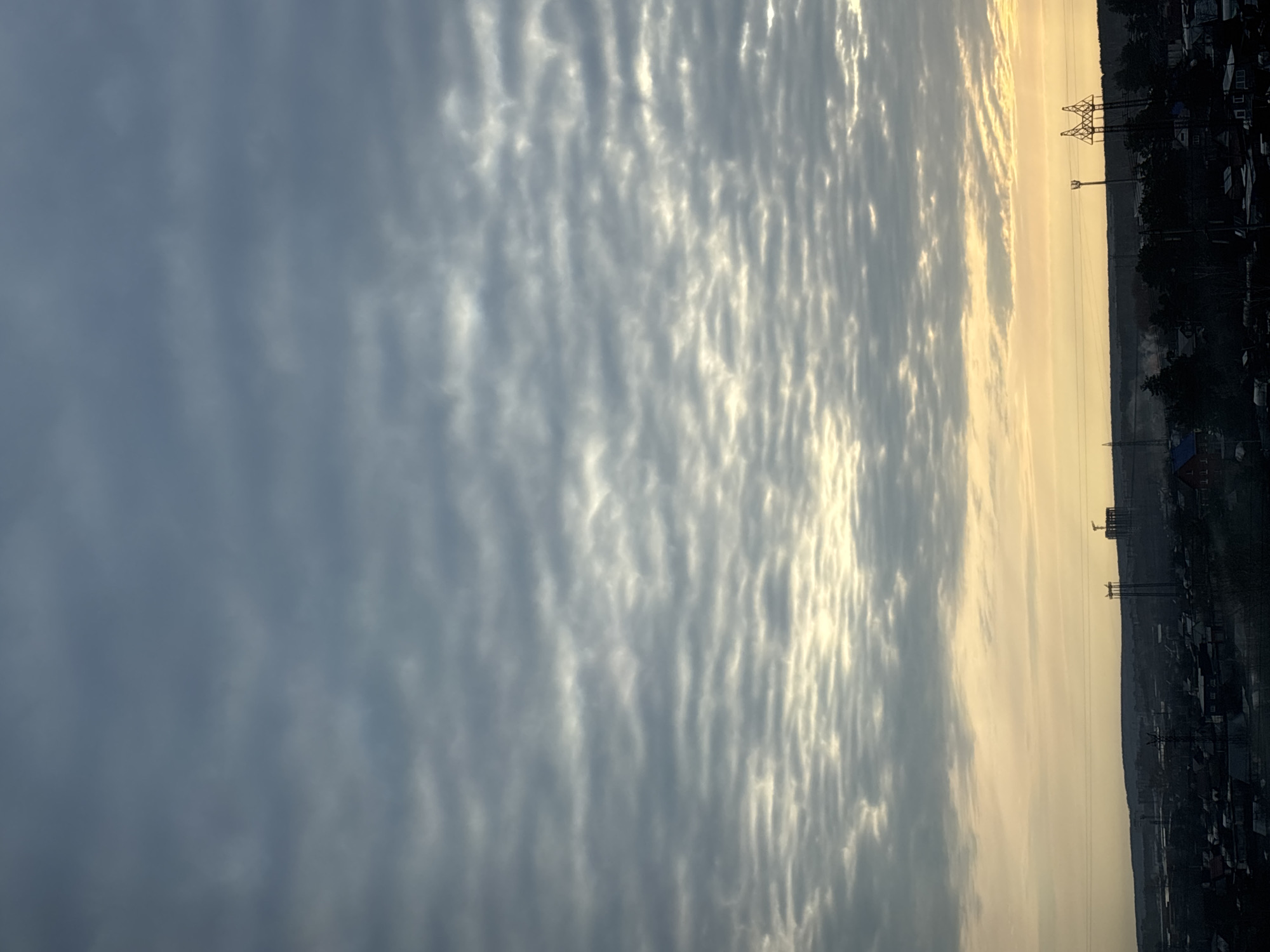 The image depicts a sky filled with striated clouds, transitioning from a darker gray to a lighter, golden hue. The sky appears to be situated over a townscape, possibly the city of Chaikovsky, Russia, based on the presence of power lines, houses, and trees visible in the background. The power lines are supported by towers, and the townscape appears somewhat silhouetted against the brighter part of the sky.