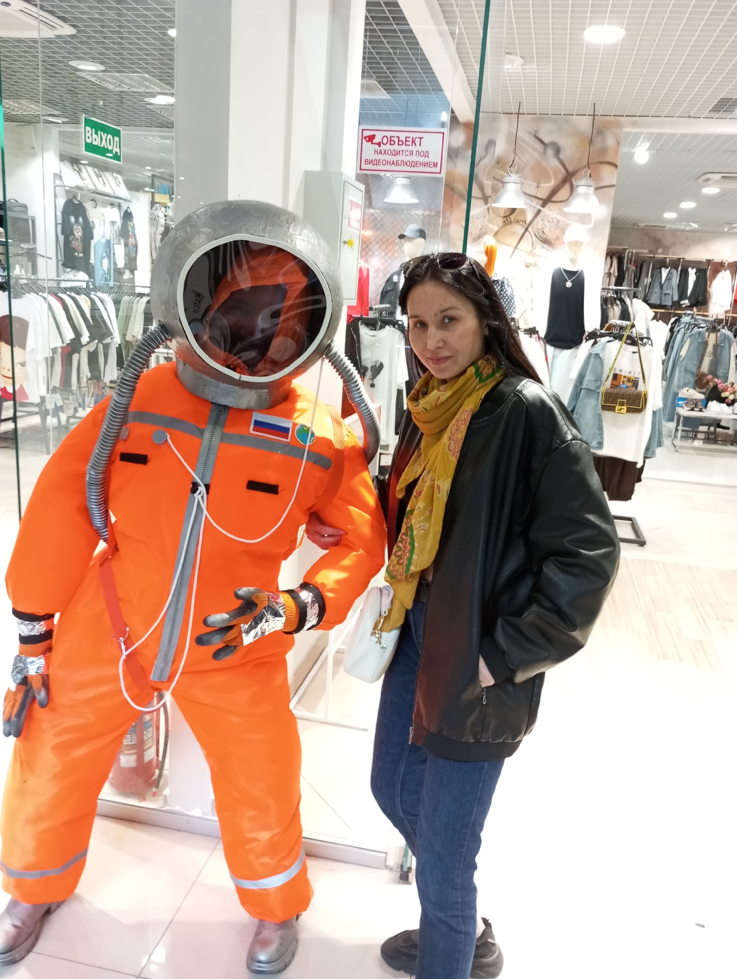 A woman poses with a mannequin dressed in an orange spacesuit with a silver helmet. The woman, wearing jeans, a black leather jacket, and a yellow scarf, is slightly behind the mannequin. The mannequin stands near a storefront window displaying various clothing items. A red fire extinguisher is visible behind the mannequin. The scene appears to be inside a shopping mall, based on the store displays. The spacesuit has a Russian flag patch and other details suggesting a space exploration theme. A sign with Russian text is visible, likely indicating the location in Izhevsk, Russia, where the photo was taken.