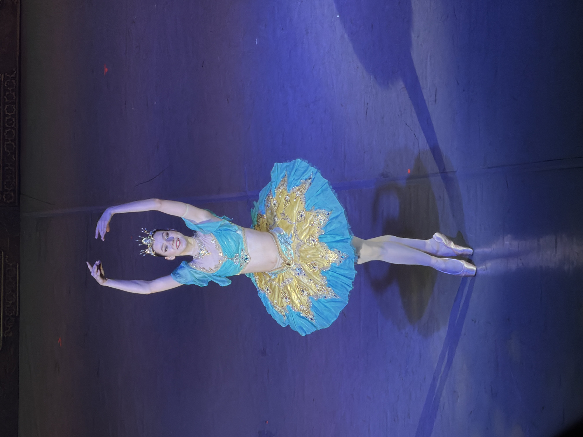 A ballerina in a light blue and gold tutu is performing on a stage. The stage is dimly lit with blue light, casting shadows on the surface. The ballerina is wearing a tiara and jewelry, and her arms are extended. The scene appears to be a ballet performance, possibly taking place in Izhevsk, Russia.