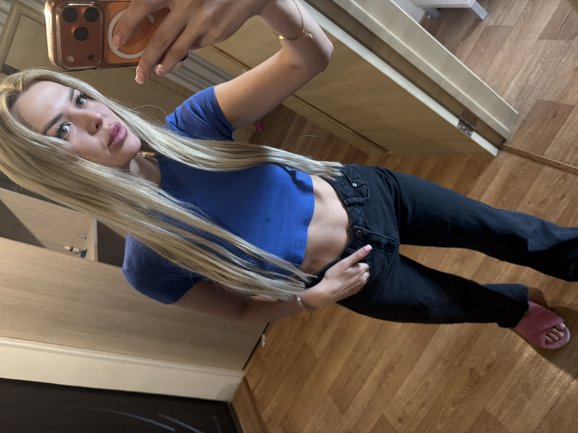 A young woman with long blonde hair is taking a selfie in what appears to be an apartment hallway. She is wearing a blue cropped t-shirt and black jeans, with one hand in her pocket and the other holding an orange phone. She has a pink slipper on her left foot. The floor is made of light brown wood. The background includes a wooden door and a light-colored wall. There are no discernible landmarks to indicate the specific location within Kyiv, Ukraine.