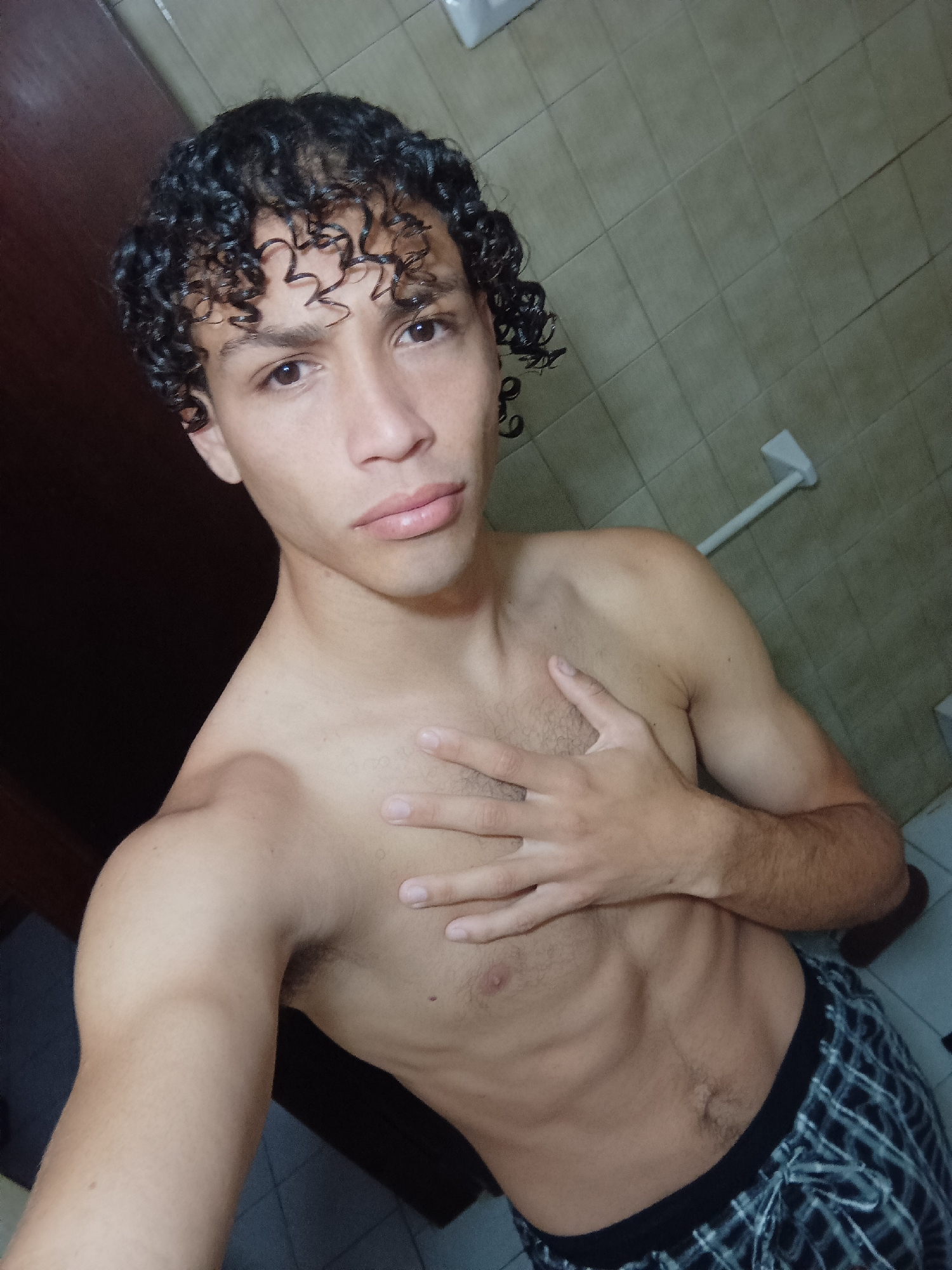 A young man with dark, curly hair and light brown eyes poses for a selfie indoors, likely in a bathroom in Porlamar, Venezuela. He is shirtless, displaying a slim physique with some visible abdominal definition and light chest hair. His left hand is placed over his chest. He wears plaid sleep shorts/boxers. His expression is neutral, looking directly at the camera.
The background consists of light brown or beige square-tiled walls. On the right side of the image, a white towel bar or rack is affixed to the wall. To the left, a dark brown wall or door frame is visible. The lighting is somewhat dim, suggesting artificial indoor lighting. No text is visible in the image.