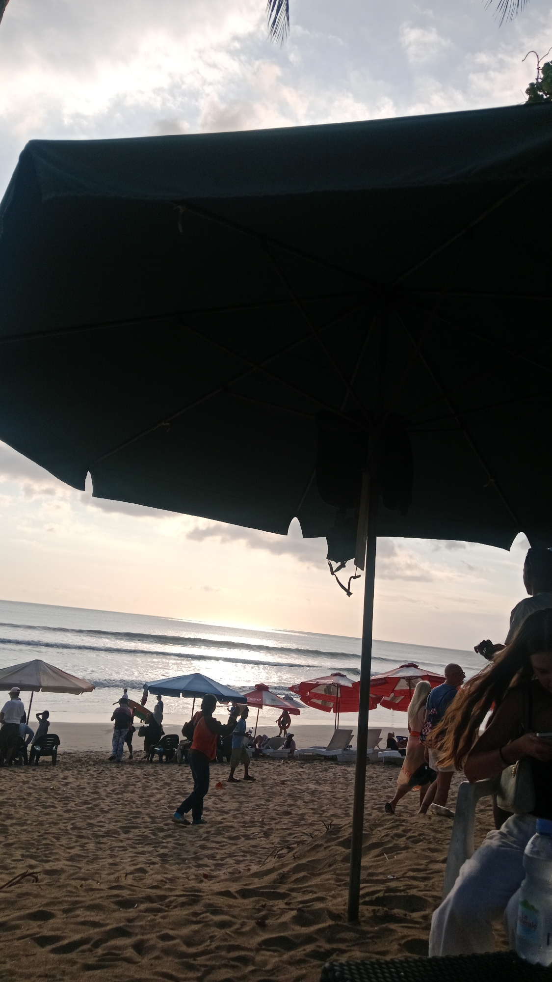 This image captures a beach scene in Kuta, Indonesia, likely during the late afternoon or early evening, as indicated by the soft lighting and the sun's reflection on the water. The foreground is dominated by a large, dark umbrella providing shade. In the midground, the sandy beach is populated with several beach umbrellas of various colors, including white, blue, and red, with some beach chairs arranged beneath them. People are visible, some walking on the sand, others appearing to be sitting or standing near the umbrellas. The ocean stretches into the background, with gentle waves rolling onto the shore. The sky is partly cloudy, with the sun casting a warm glow. The overall atmosphere suggests a relaxed beach setting with people enjoying the seaside.