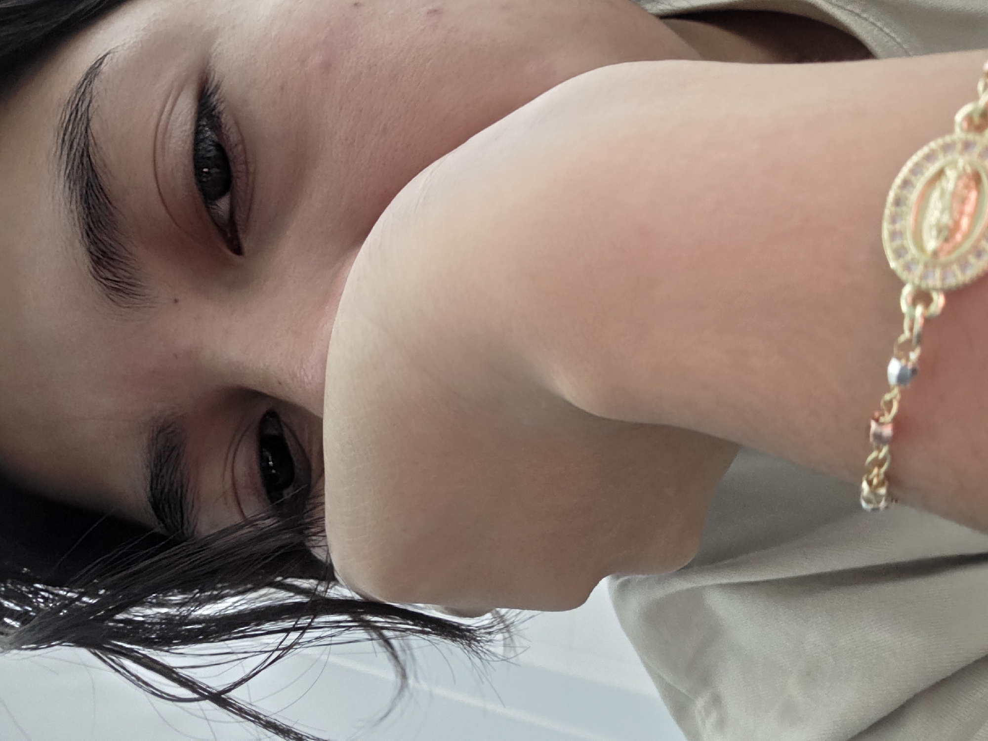 This is a close-up, first-person perspective photograph of a person's face and arm, taken in La Tijera, Mexico. The subject's face is partially obscured by their right hand, which is resting against their mouth. Dark hair frames the left side of the face. The subject is wearing a light-colored t-shirt. A gold bracelet with a religious medallion, appearing to depict a figure, is visible on the subject's wrist. The background is a plain, light-colored wall.