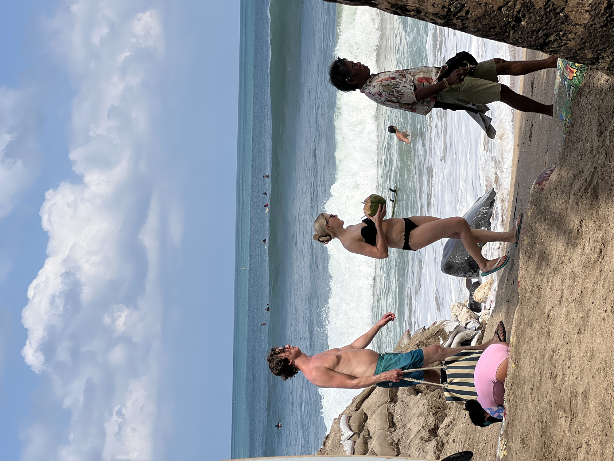 In Kuta, Bali, Indonesia, a group of people are on the beach near the ocean. In the foreground, a man with brown hair and glasses walks to the left, wearing blue swim trunks and carrying a striped beach bag. Beside him, a woman with blonde hair holds a coconut and wears a black bikini. To the right, a man with dark hair wears a colorful shirt and khaki shorts. Further back, a child in a pink swimsuit lies on the sand, and a person in the ocean swims. Surfers are visible on the water in the distance. The beach is sandy with some rocks and sandbags near the shore. The ocean has waves and a clear blue sky with clouds overhead.