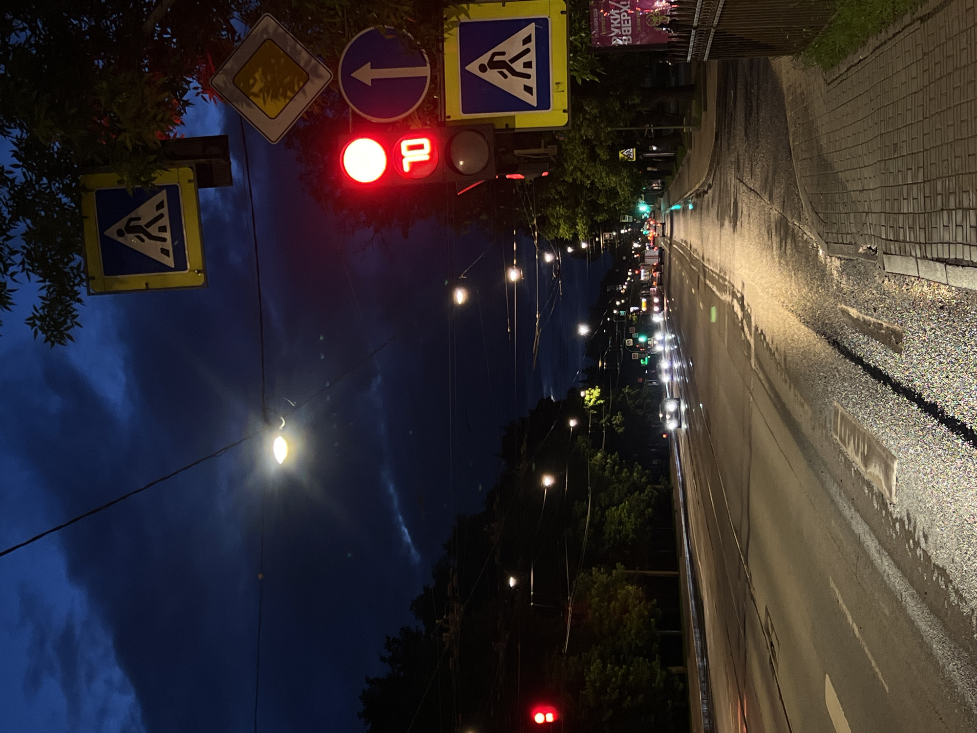 The image depicts a twilight scene on a city street in Saint Petersburg, Russia, likely during or after rain, as the asphalt is wet and reflective. A traffic light is visible, showing a red light and a digital display indicating "10". There are multiple traffic signs: a pedestrian crossing sign (with a blue background and white pedestrian silhouette) and a warning sign with a diamond shape and a yellow background. Several vehicles are present on the road, their headlights illuminated. Trees line the street, and overhead wires are strung across the scene, consistent with street lighting and potentially trolleybus power lines common in Saint Petersburg. The sky is dark blue with dramatic clouds. The overall atmosphere is urban and moody, illuminated by artificial lights and the fading natural light.