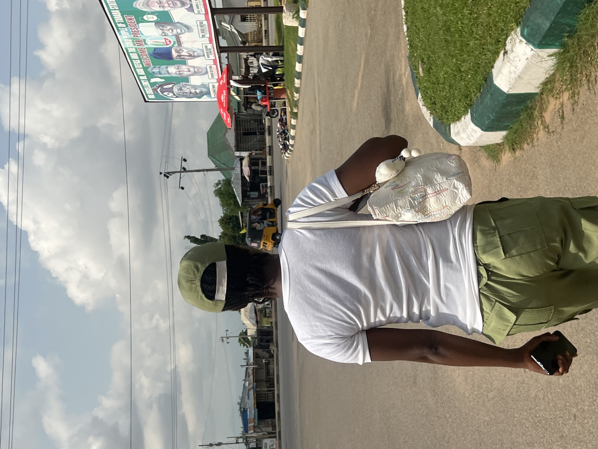 This media file shows a person from behind, wearing a white t-shirt and green uniform pants, and a green cap. They are holding a white bag and a mobile phone. The person appears to be outdoors, possibly on a street or pathway. In the background, there is a billboard with images of people and text that reads "WELCOME MR. PRESIDENT" and "APC". There are also some buildings, trees, and power lines visible. The sky is partly cloudy. The scene suggests a public gathering or event, possibly related to a political campaign. The presence of a billboard welcoming the president and the "APC" branding points to a political context in Nigeria, specifically in Jalingo. The image is taken during the daytime, with good lighting. The overall mood is neutral, with no clear emotional expressions visible from the subject.