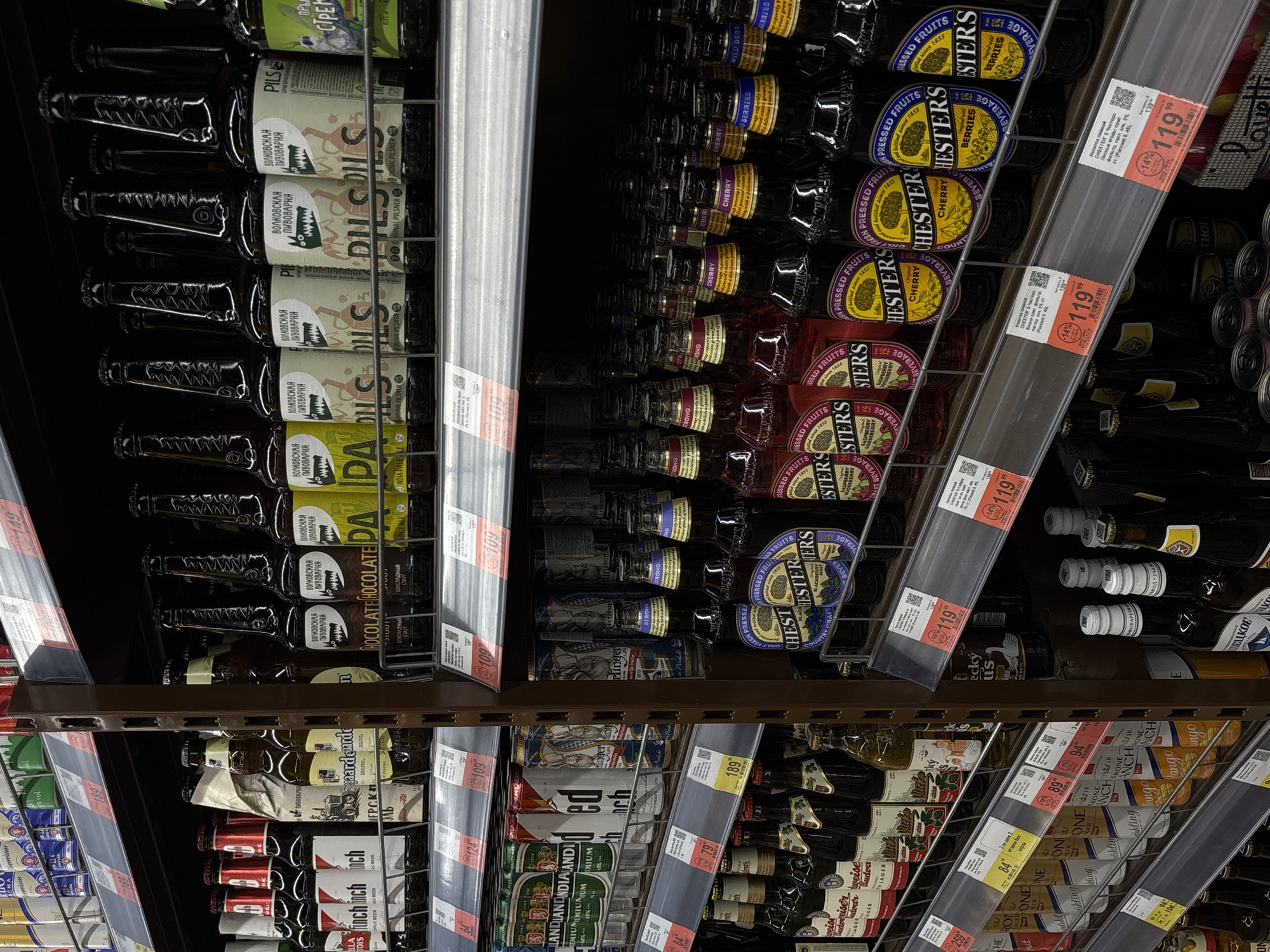The image shows a supermarket shelf in Perm, Russia, stocked with various alcoholic beverages. The shelves are dark brown and metal, with transparent price tags attached to the front. A variety of beers and other alcoholic beverages, primarily in glass bottles, are arranged in rows. Brands like "Волковская Пивоварня" are visible, alongside fruit-flavored drinks labeled "CHESTER’S" with flavors like cherry and berries. There are promotional price tags indicating prices around 119 and 189 rubles. The scene is brightly lit and suggests a typical retail environment.