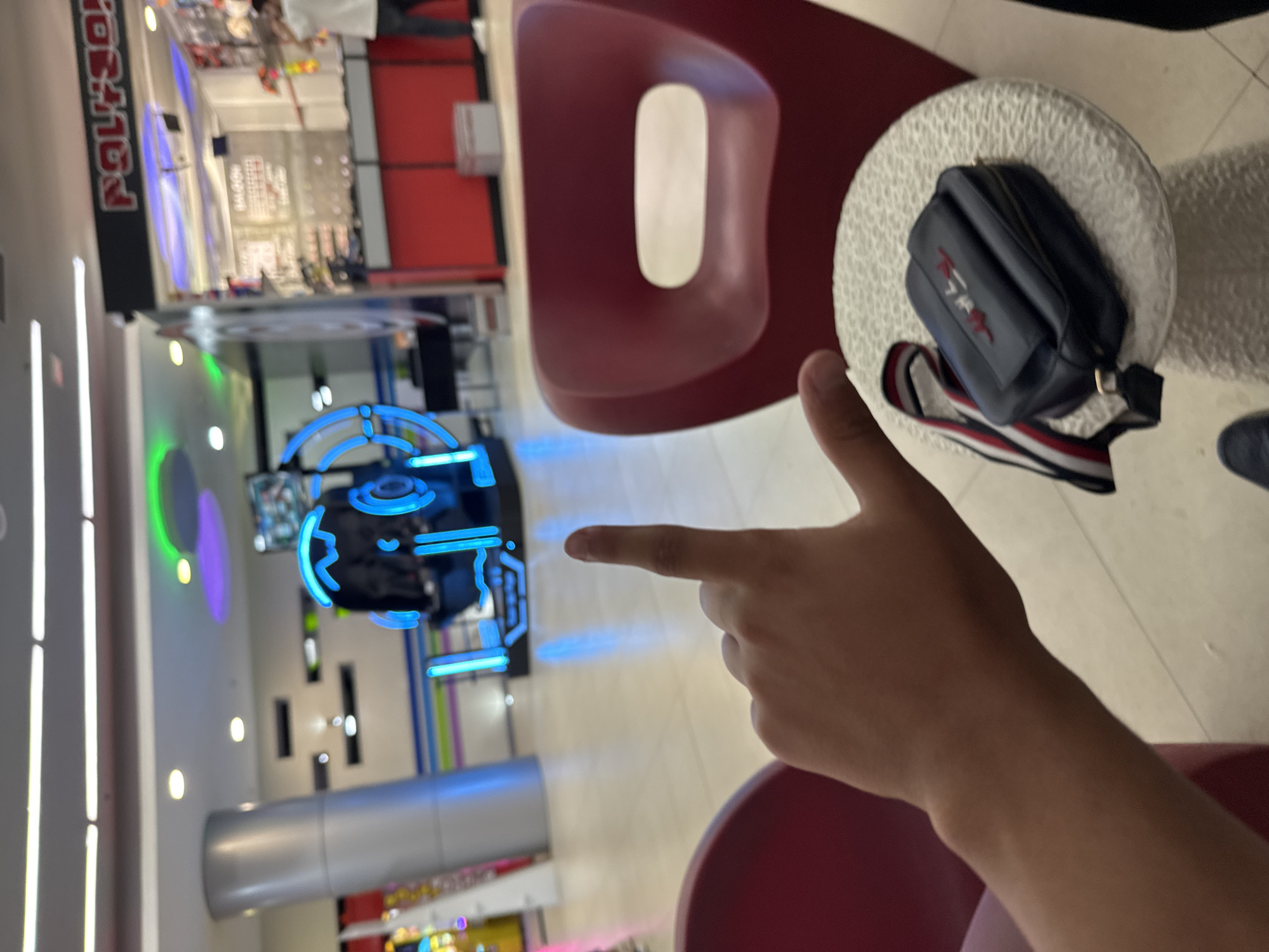 An indoor photograph, likely taken in a shopping mall or entertainment center in Yerevan, Armenia. In the immediate foreground, a person's right hand and forearm are visible, with the index finger extended and pointing towards a brightly lit arcade game or entertainment console in the middle-ground. The console features prominent blue neon-like lighting, outlining its structure and reflecting on the polished floor.
To the right, a dark red, modern-styled armchair with a distinctive oval cut-out in its backrest occupies part of the frame. Adjacent to the armchair, on a small, textured white cylindrical stool or table, rests a dark blue shoulder bag. The bag has a strap with red, white, and blue stripes, and an embroidered logo in red and white that reads "TOMMY" (Tommy Hilfiger).
The background features a bustling indoor environment with light-colored tiled flooring and various artificial lights illuminating the space. Reflections on polished surfaces suggest glass storefronts or displays. A large red sign with blurry, unidentifiable white text, appearing inverted, is visible in the upper background, likely indicating a store or attraction name. The overall scene conveys a casual, leisure activity in a public indoor space.